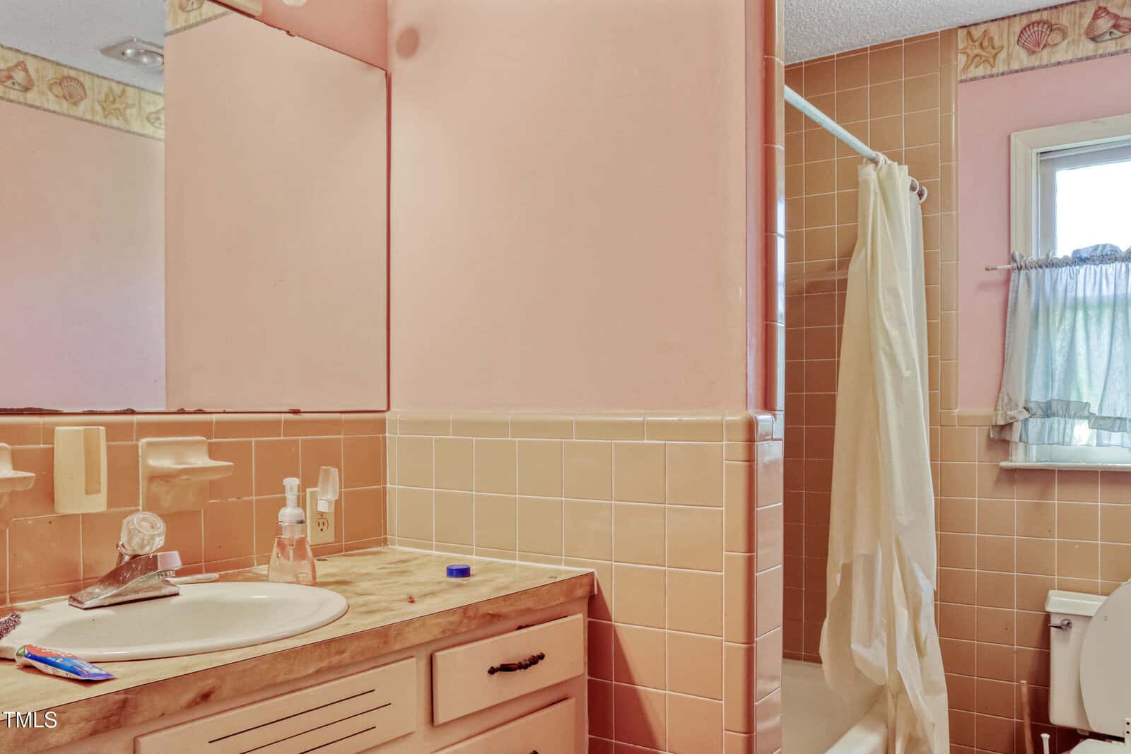 A bathroom with pink and tan tile walls features a sink with a marble-patterned countertop, a mirror above it, and a toothbrush and soap dispenser. A walk-in shower with a white curtain is on the right, next to a window with a blue curtain and a toilet.