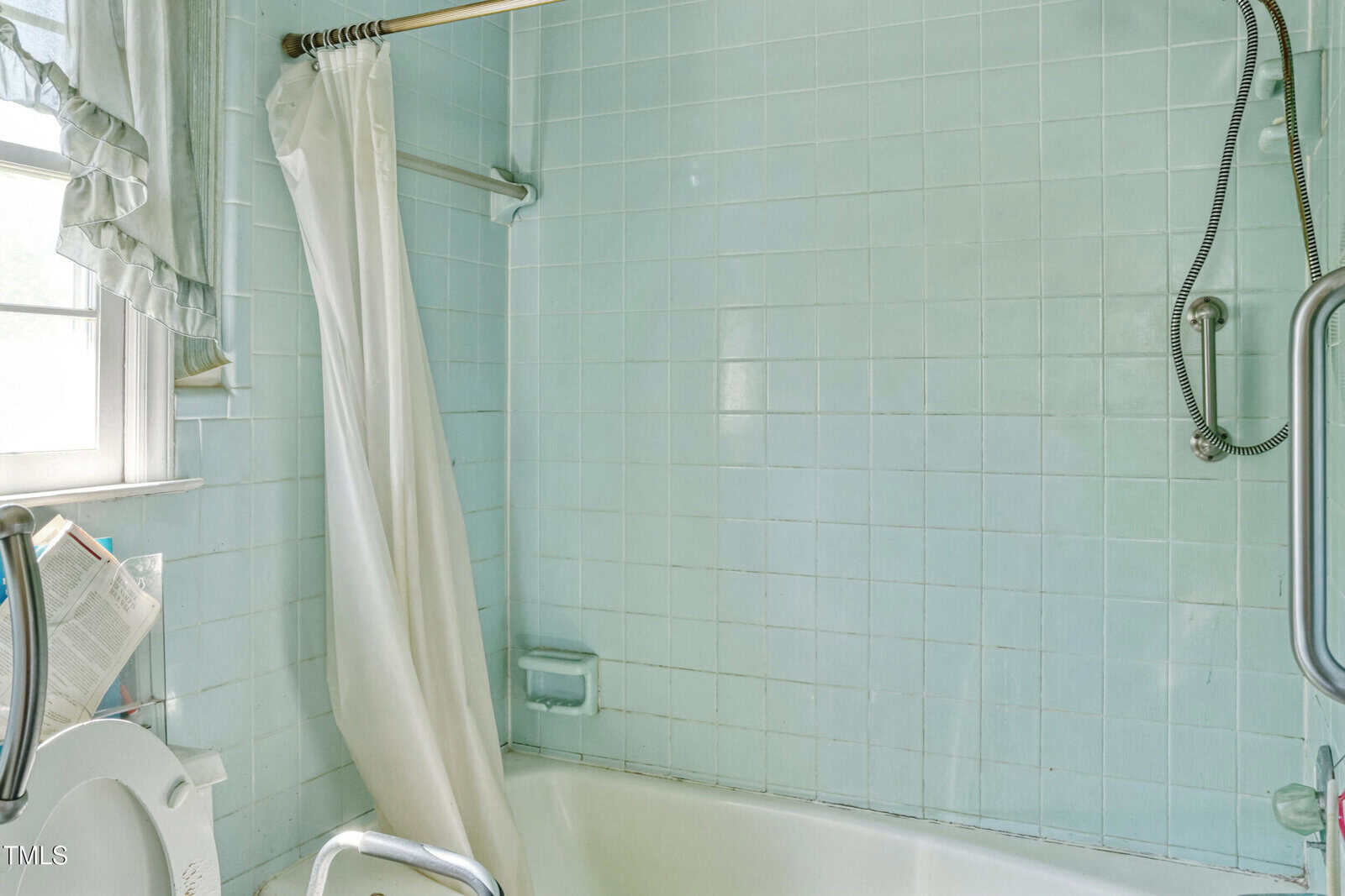 A bathroom with light blue tiles features a bathtub with a white curtain partially drawn across. A window with sheer curtains is on the left, and there is a metal grab bar next to the tub. A showerhead is visible above the bathtub.