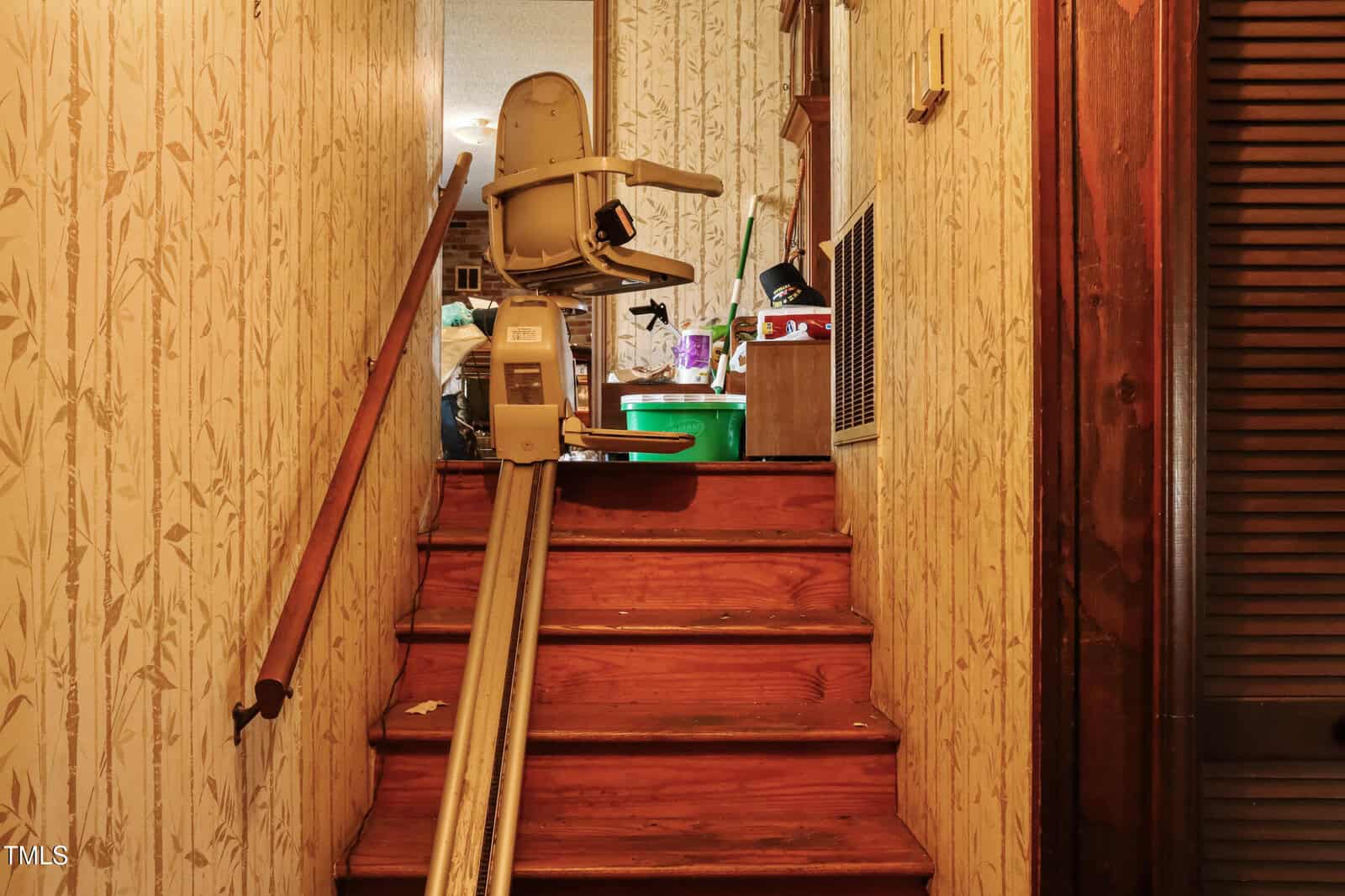 A wooden staircase with a stair lift installed on the right side. The surrounding walls have floral-patterned wallpaper. At the top, various boxes and a green bucket are visible in a cluttered hallway.