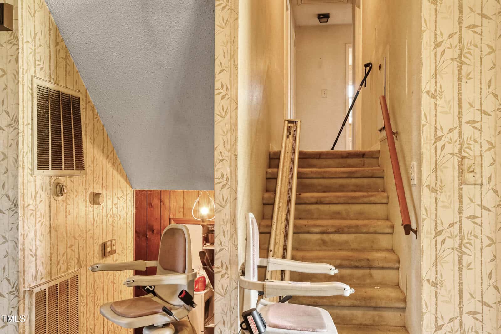 A stairwell with beige floral wallpaper features a chair lift on the left side. The stairs are carpeted in a light brown color and lead to a well-lit upper hallway. Wooden handrails are present on the right side of the stairs.