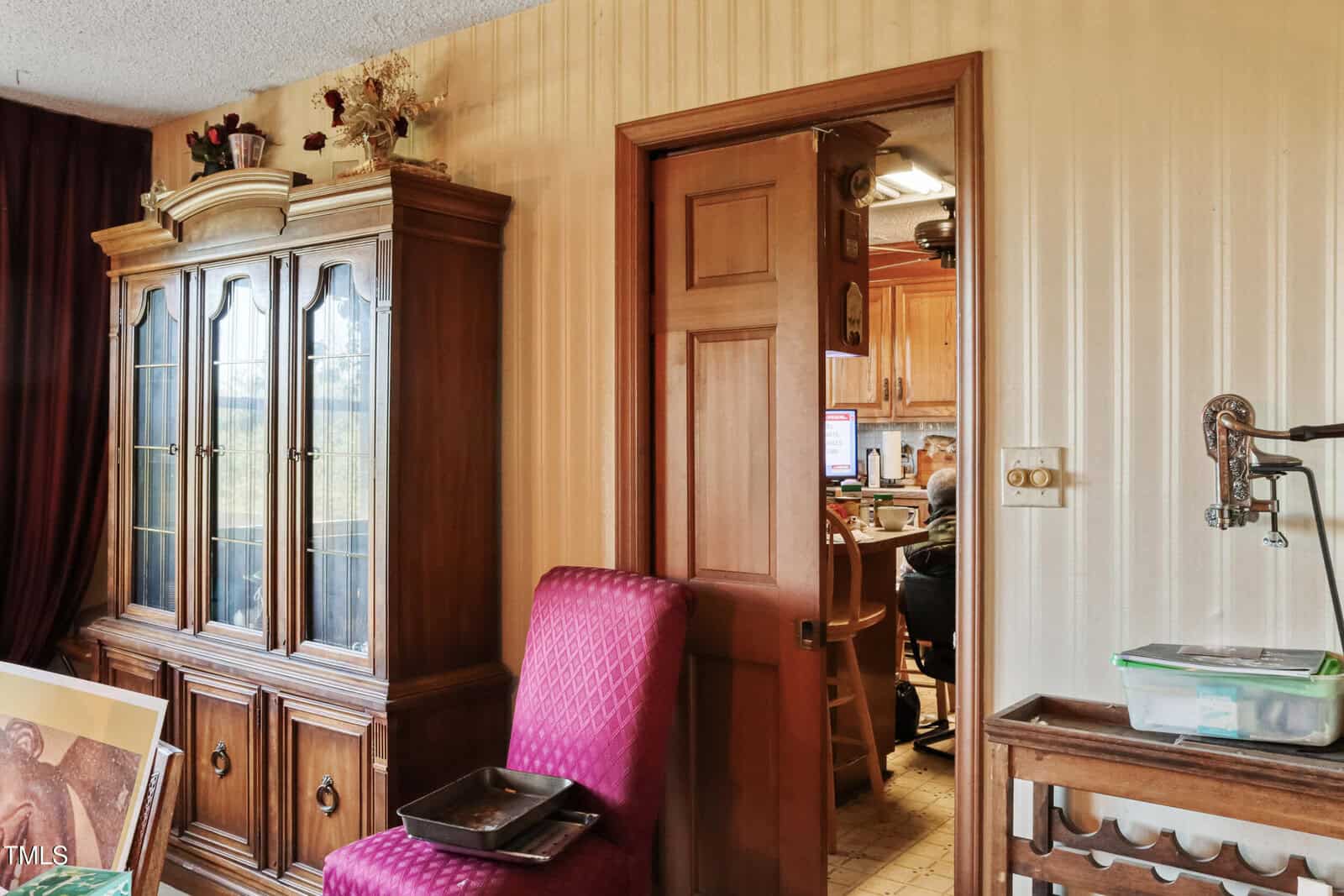 A room with a wooden china cabinet on the left, featuring glass doors and decorative items on top. A bright pink upholstered chair is in front of it. A partially open door leads to a kitchen with a person sitting at a computer. An antique faucet is visible on the right.