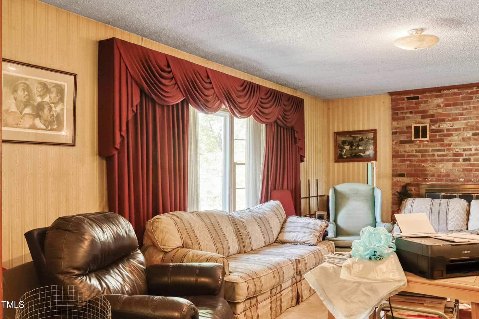 A living room with a striped couch, leather recliner, and wooden paneling. Large draped curtains flank a window. A brick wall is partially visible. A framed image and assorted furniture complete the space.