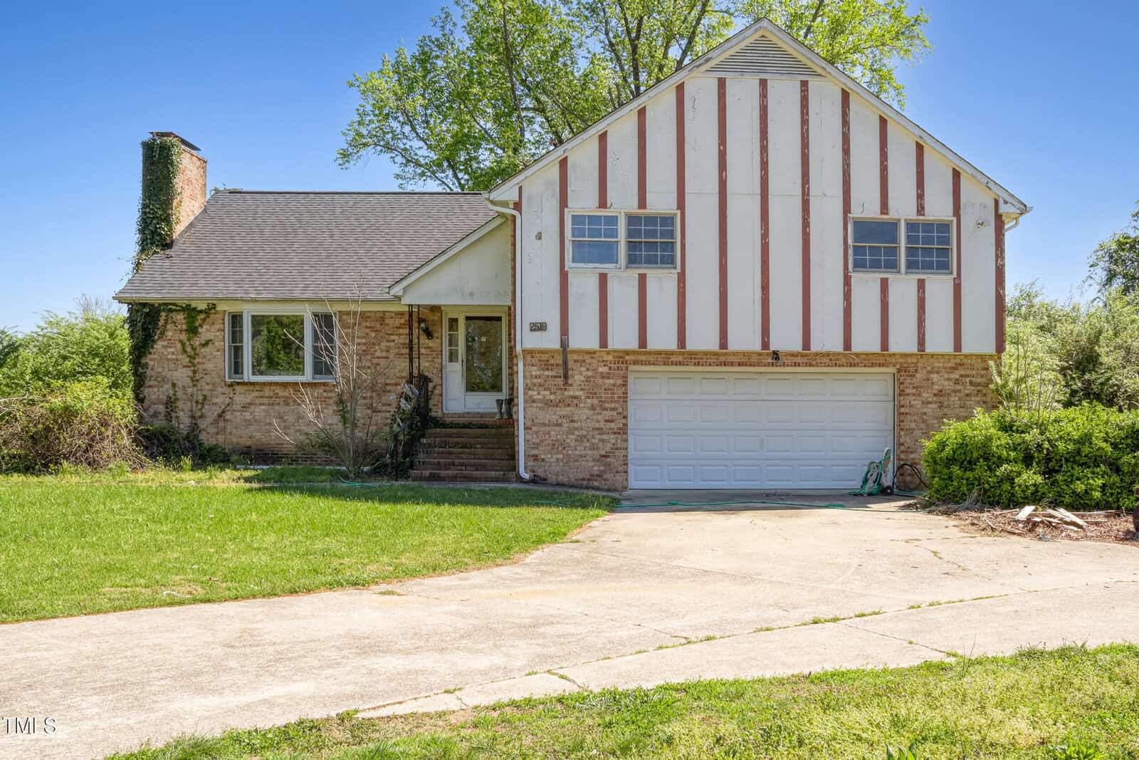 Nestled under a clear blue sky at 2510 Poplar Creek Road, this split-level house in Henderson boasts a charming brick exterior with white paneling. It features a double garage, an inclined driveway, and an ivy-covered chimney. The yard is mostly grass, with shrubs framing the entrance steps.
