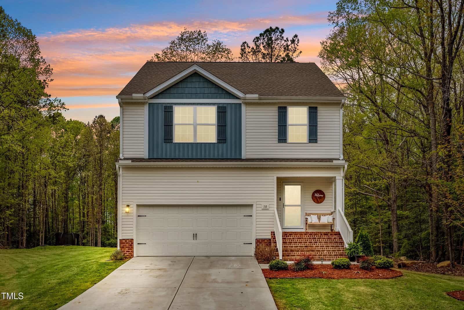 Two-story house with white siding and blue shutters, a double garage, brick accents, and a concrete driveway, surrounded by trees and grass under a colorful sunset sky.