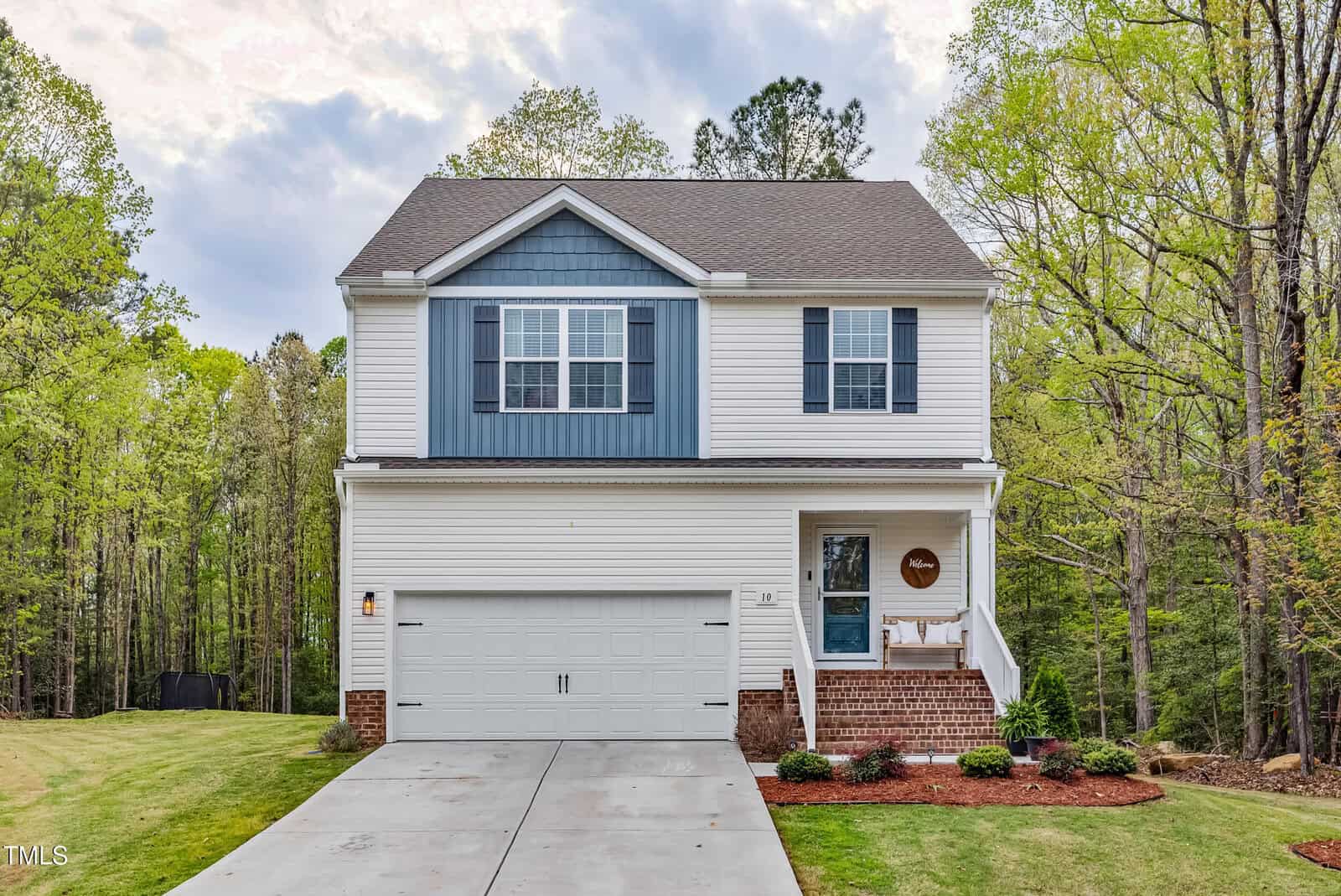 Two-story house with white siding, blue accent panels, and a brick foundation. There is a double garage, a small front porch with a bench, and a concrete driveway. The home is surrounded by grass and trees.