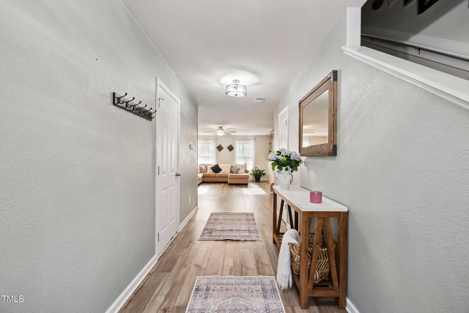 A hallway with light gray walls, wood flooring, and two rugs leads to a living room. On the right, a wooden console table holds decor and a mirror hangs above. A coat rack is mounted on the left wall.