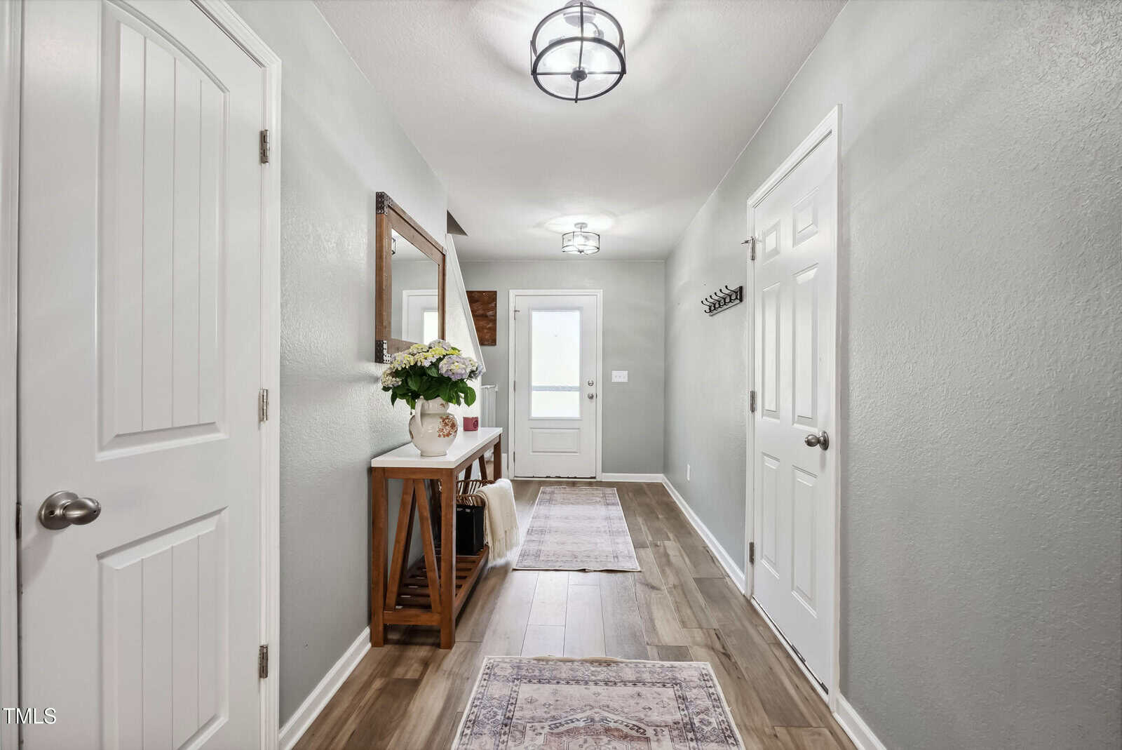 A hallway with light gray walls, wood-look flooring, and two ceiling lights. A wooden console table with a flower arrangement and mirror is on the left, and two rugs are laid along the floor. Several closed doors are visible.