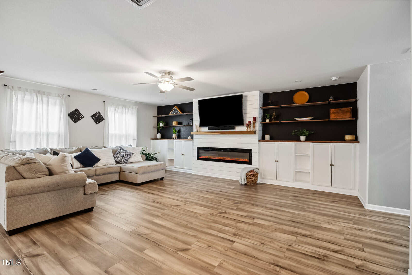 A spacious living room with wood flooring, a beige sectional sofa, a ceiling fan, built-in shelves, a wall-mounted TV above a fireplace, and large windows with white curtains.