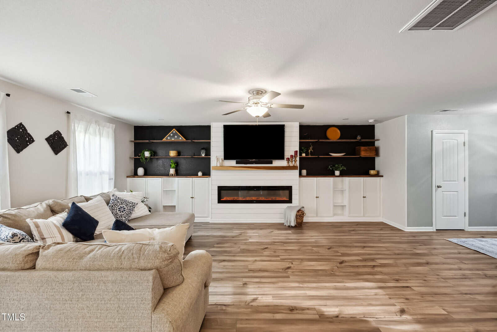 Spacious living room with a beige sectional sofa, wood flooring, built-in shelves with decor, electric fireplace, mounted TV, ceiling fan, and large window with sheer curtains. White and black color scheme.
