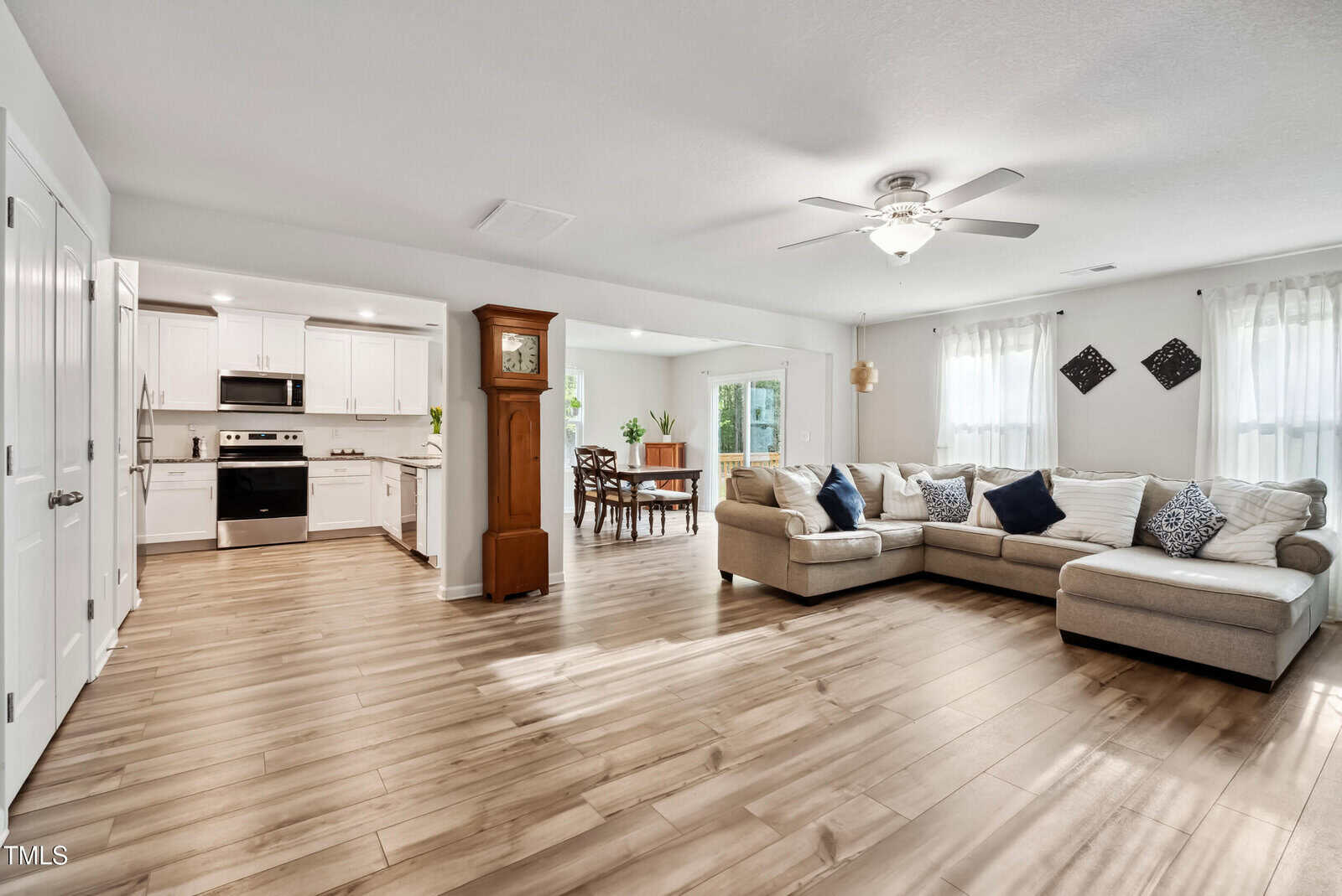 Open living space with light wood floors, a beige sectional sofa, ceiling fan, and tall clock. Kitchen with white cabinets and stainless steel appliances is visible, along with a dining table in a bright nook.