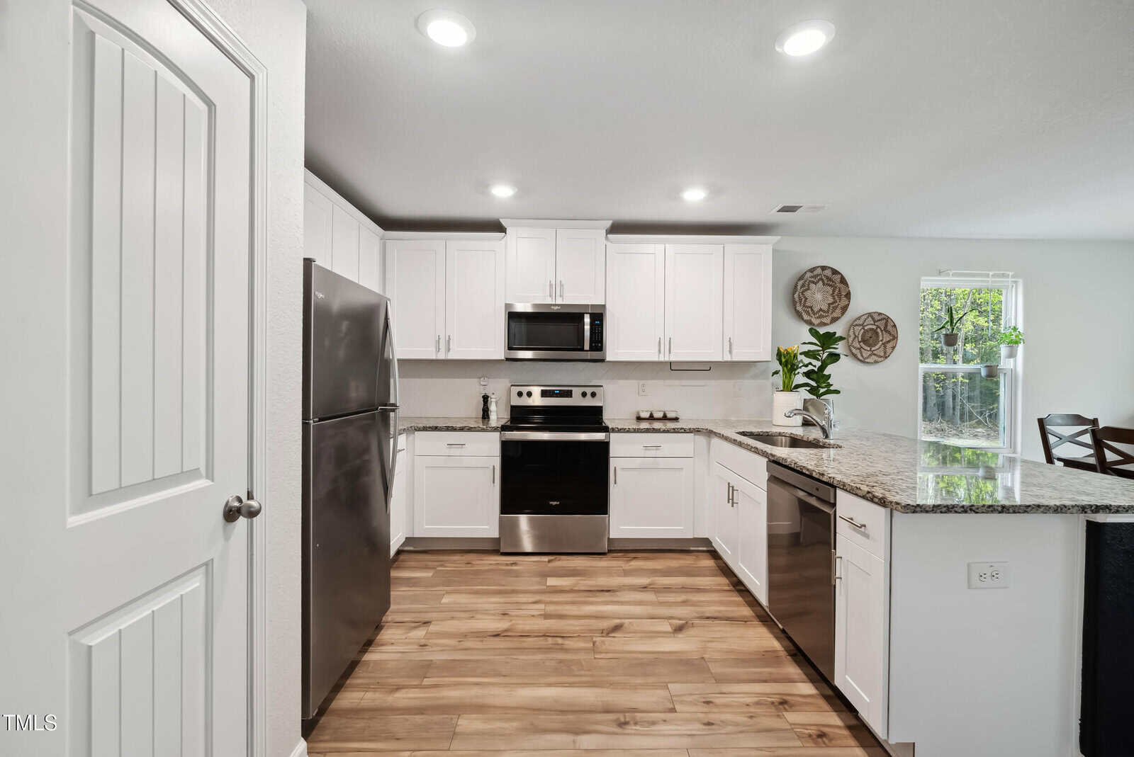 Modern kitchen with white cabinets, stainless steel appliances, granite countertops, wood flooring, recessed lighting, and a window allowing natural light. Decorative plates and a plant are on the far counter near the window.