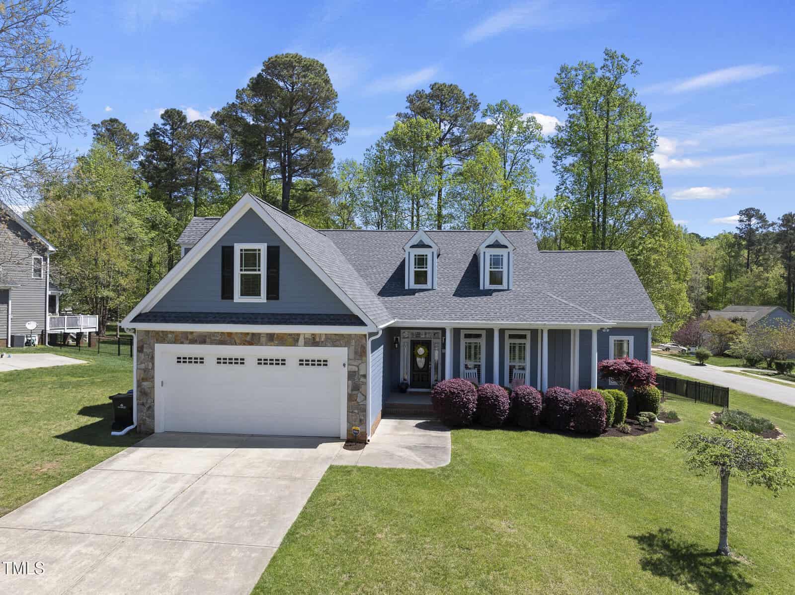 A light blue suburban house with white trim, two dormer windows, and a two-car garage. The front yard has a manicured lawn, bushes, and a concrete driveway. Tall trees and neighboring homes are visible in the background.