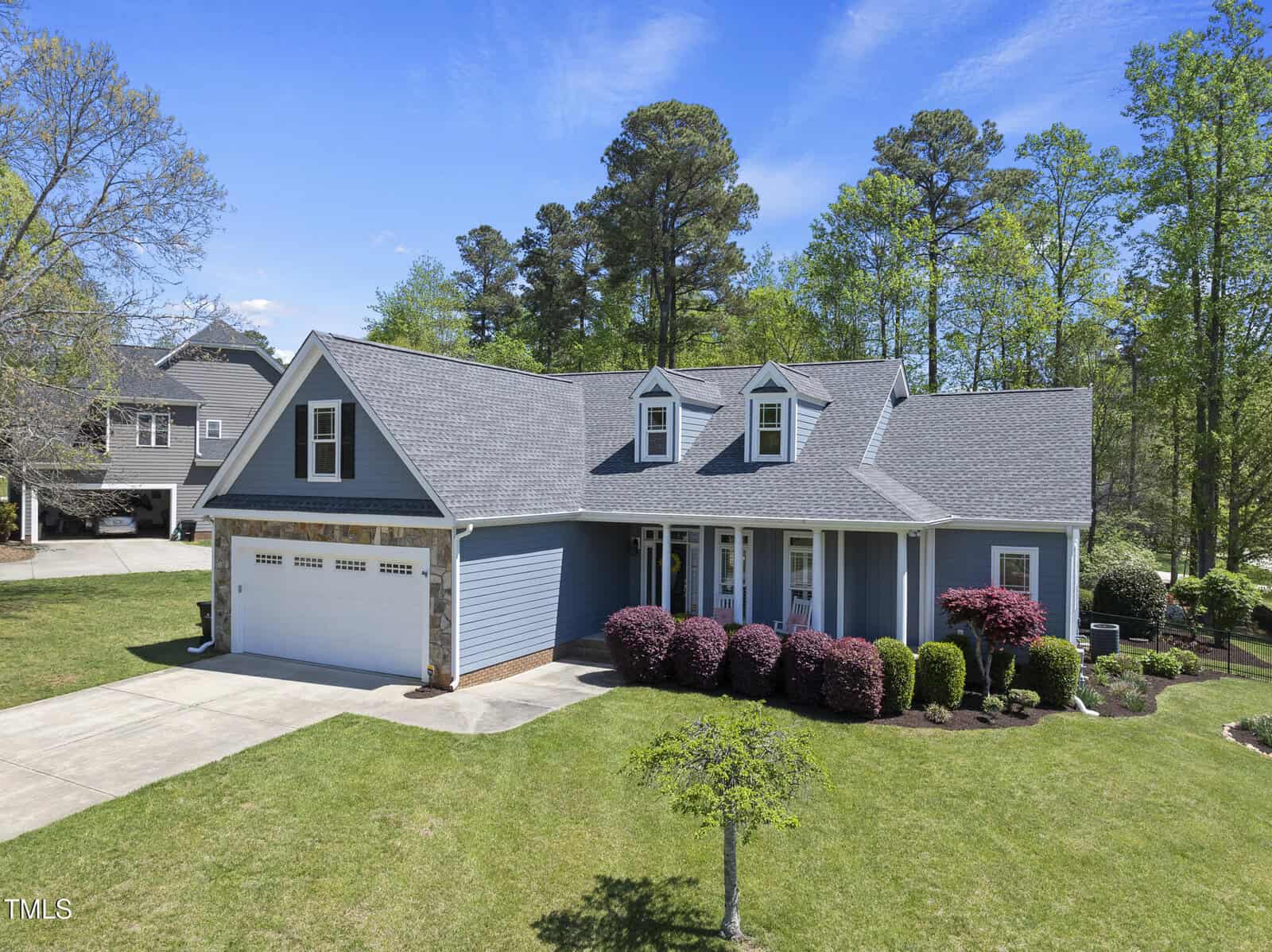 A one-story blue house with a gray shingled roof, two dormer windows, a double garage, a covered front porch, and landscaped bushes on a green lawn, with trees in the background under a blue sky.