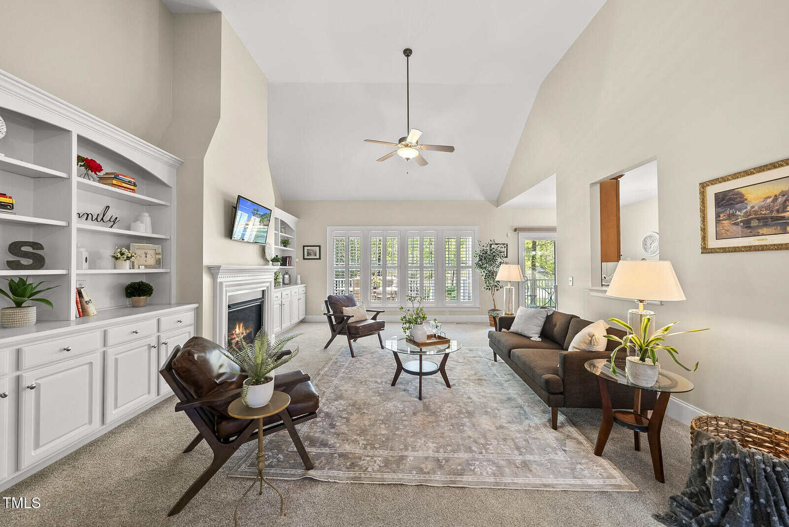 A spacious living room with high ceilings, neutral walls, and large windows. The room features a sofa, two armchairs, a fireplace, built-in shelves, indoor plants, and a rug on carpeted flooring.
