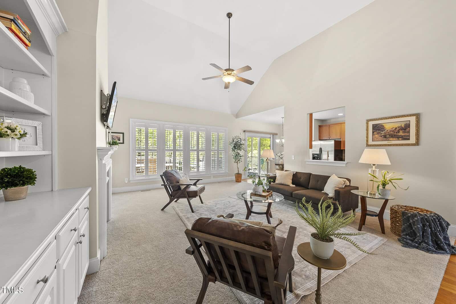 A spacious, well-lit living room with beige walls, carpeted floor, modern furniture, large windows with shutters, a ceiling fan, built-in shelving, plants, and a view into an adjacent kitchen area.