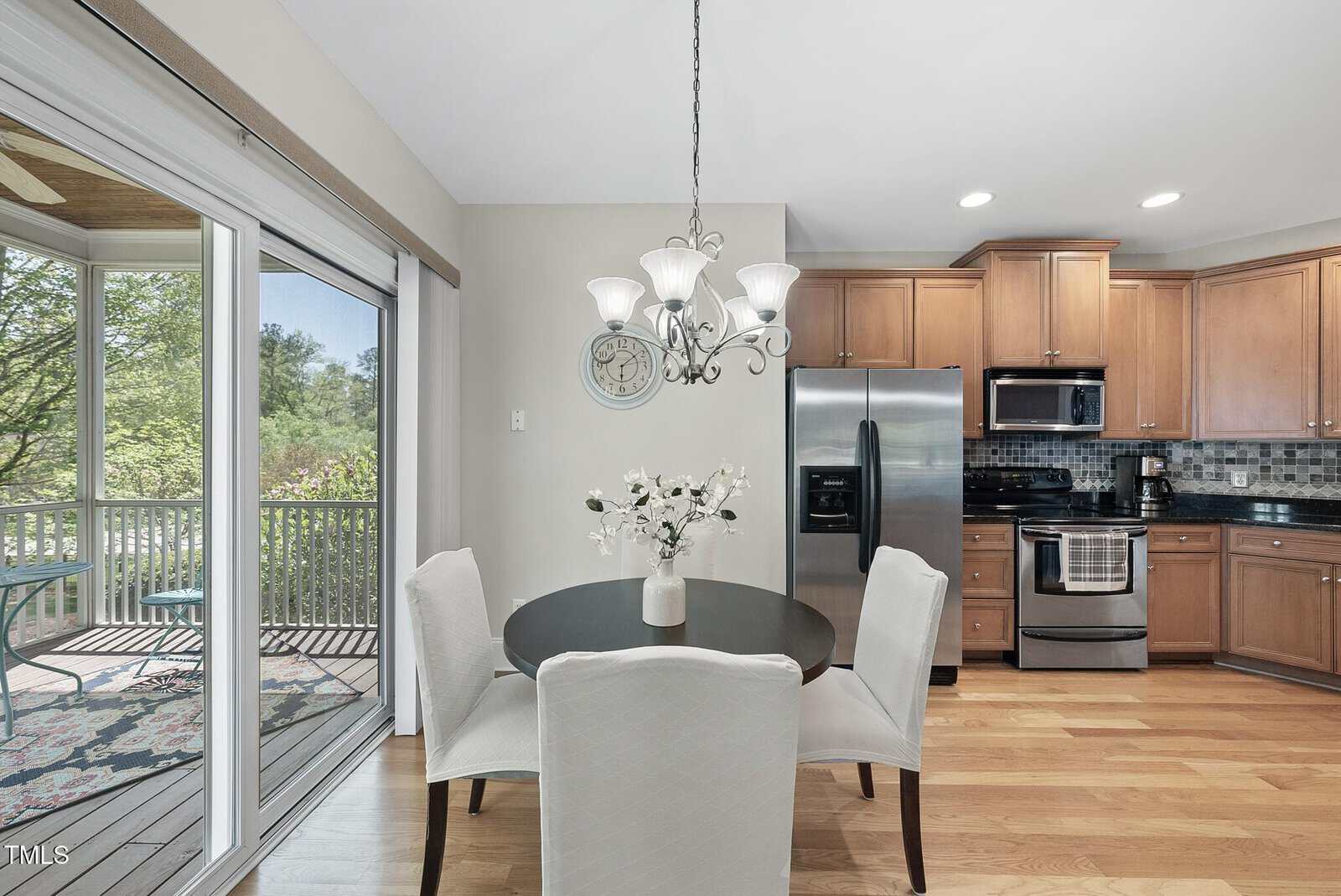 A modern kitchen and dining area with wooden floors, stainless steel appliances, a round table with four white chairs, a chandelier, and a sliding glass door leading to a porch with outdoor seating and greenery.