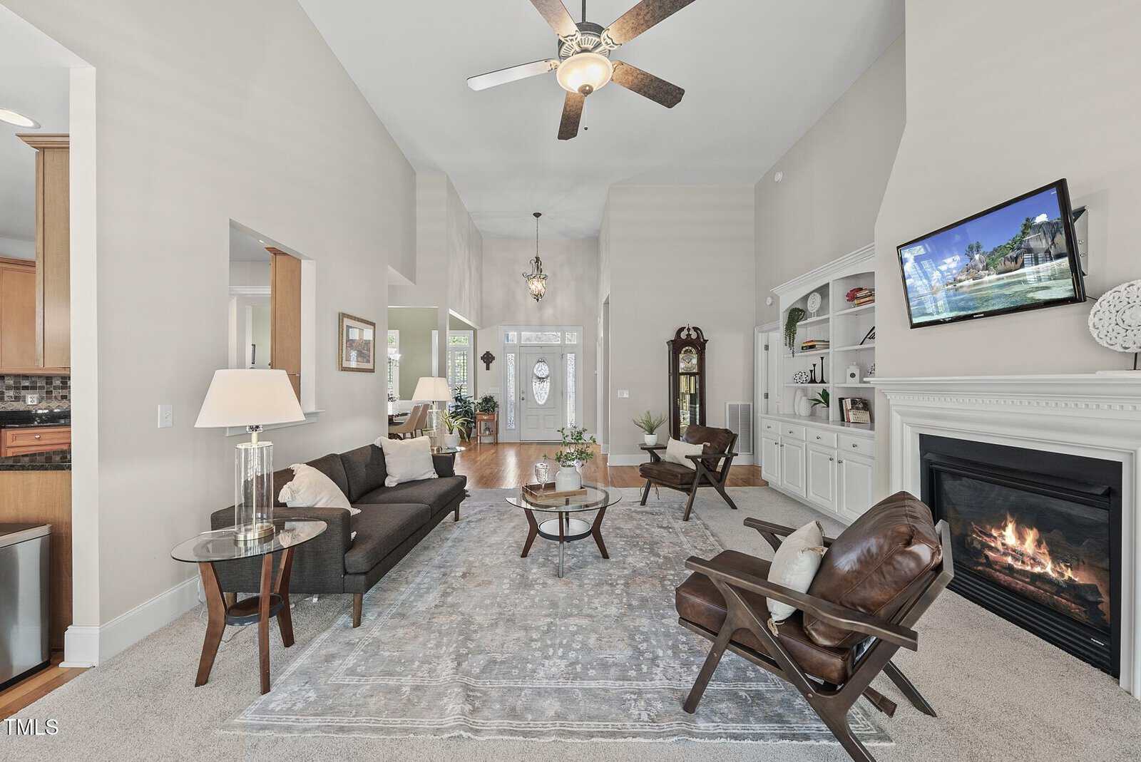 Spacious living room with neutral walls, a dark sofa, two chairs, rug, glass tables, fireplace, built-in shelves, TV above the mantel, ceiling fan, and view of the entryway and dining area in the background.