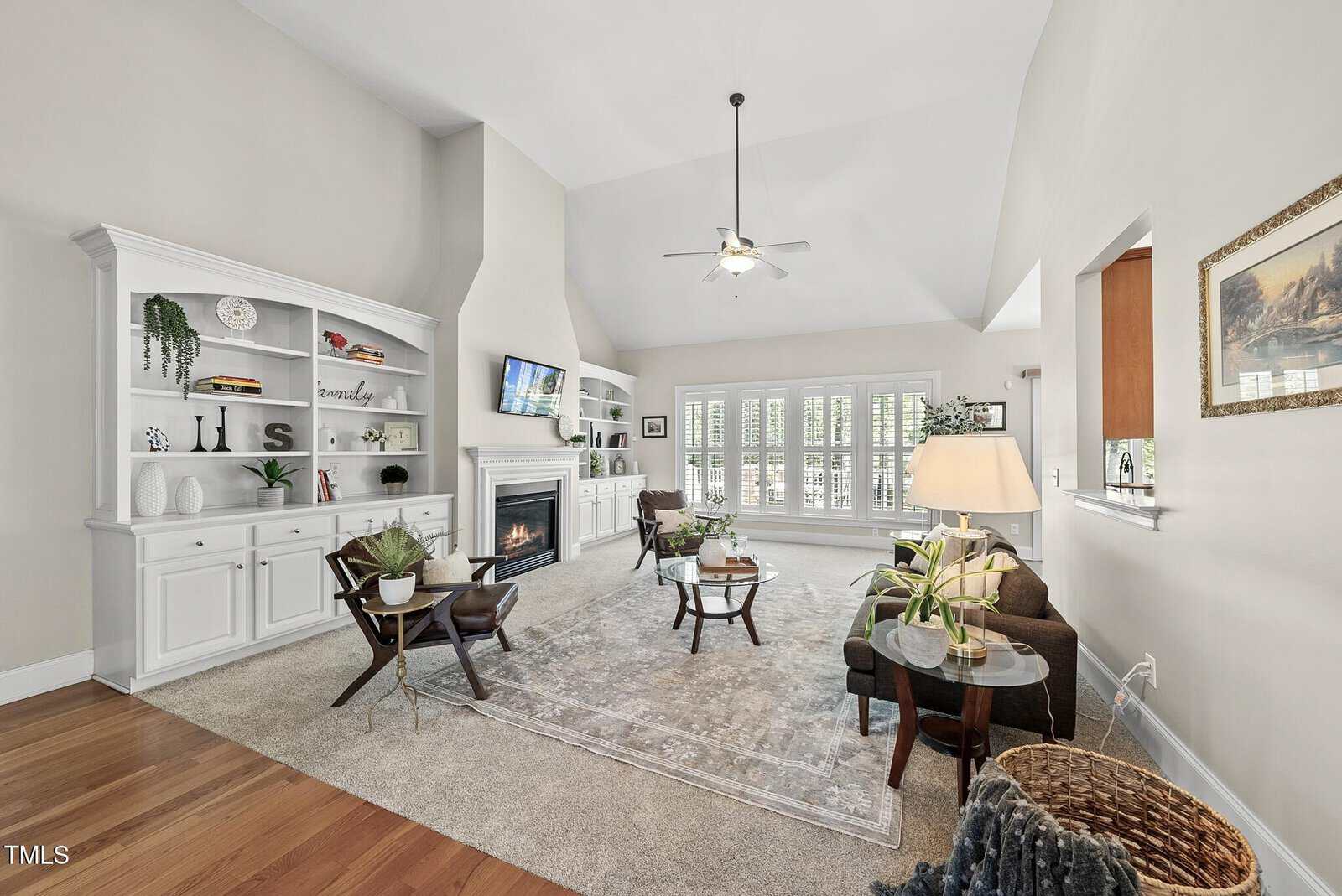 Spacious living room with vaulted ceiling, white built-in shelves, a fireplace, wall-mounted TV, neutral furniture, large windows with shutters, indoor plants, and an open view into the adjacent kitchen.