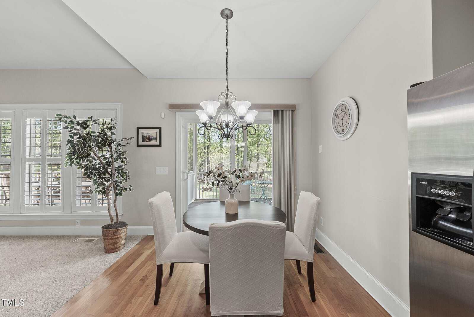 A dining area with a round table and four chairs under a chandelier, near a sliding glass door. There is a large plant, a wall clock, a stainless steel refrigerator, and hardwood flooring.