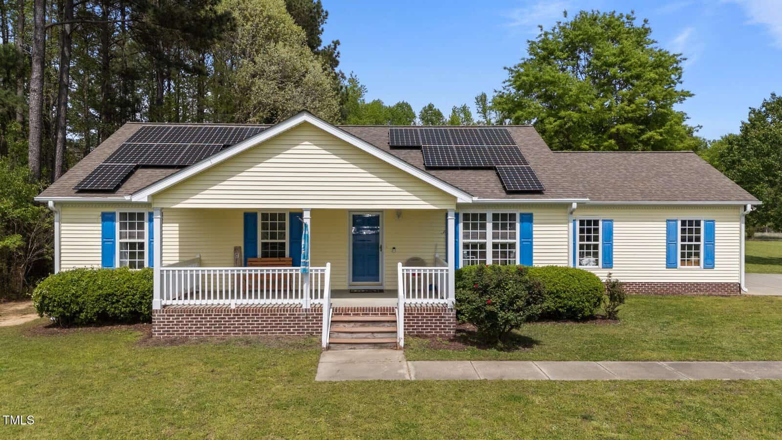 Single-story yellow house with blue shutters, a covered front porch, and solar panels on the roof. The yard is grassy with shrubs, and trees are visible in the background under a clear sky.