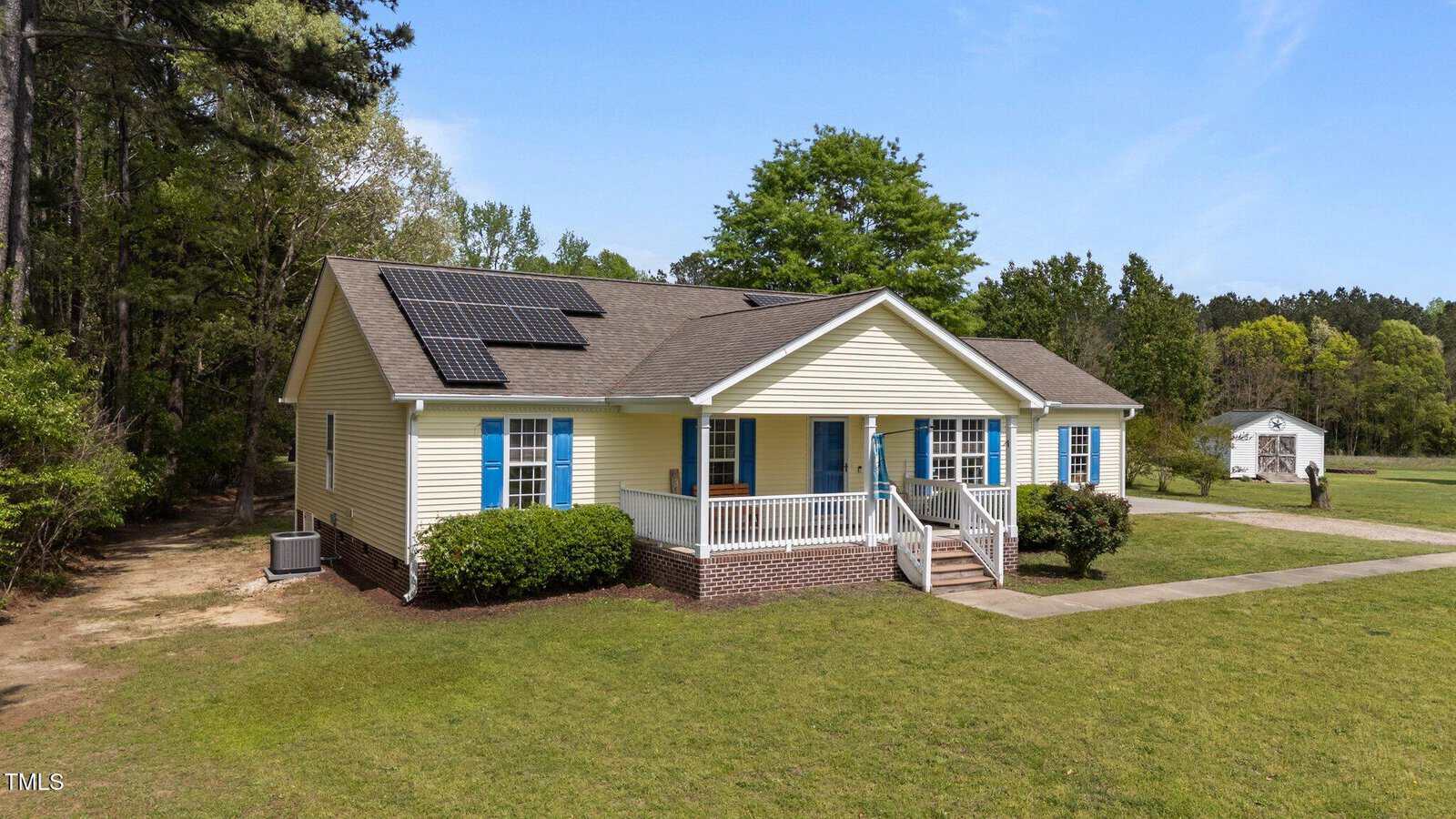 A single-story yellow house with blue shutters and solar panels on the roof, surrounded by a grassy yard, bushes, and trees. A small white shed is visible in the background.