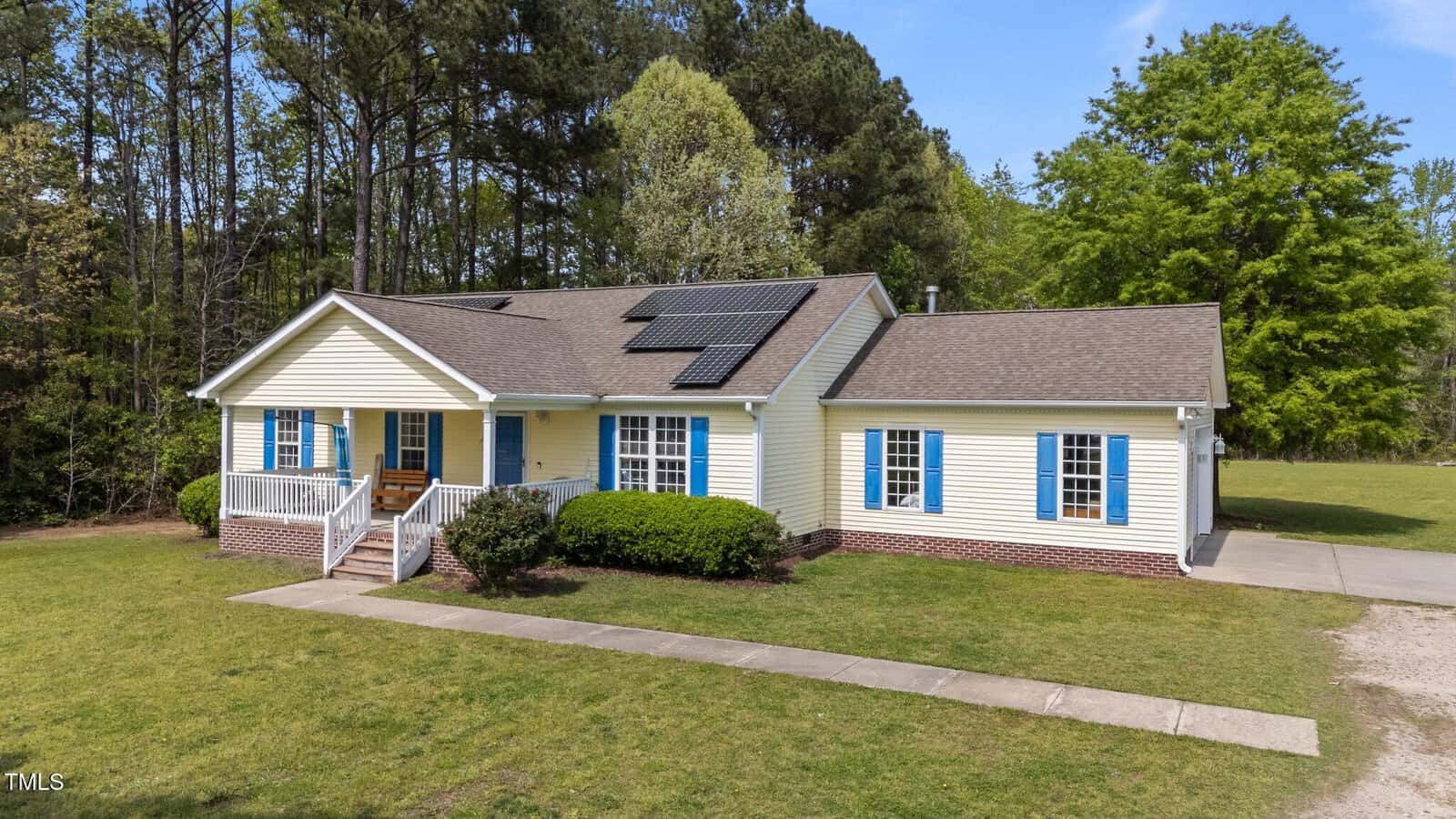 Single-story yellow house with blue shutters, white trim, and a small front porch. Solar panels are installed on the roof. The house is surrounded by grass, trees, and a concrete walkway leading to the front door.