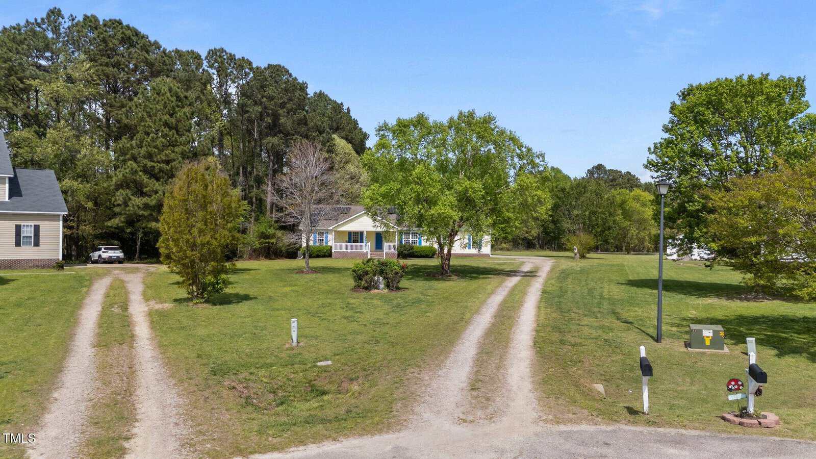A single-story house with light siding sits back from the road, surrounded by trees and grass. Two unpaved driveways lead from the street to the house. There are two mailboxes near the road and a streetlamp on the right.