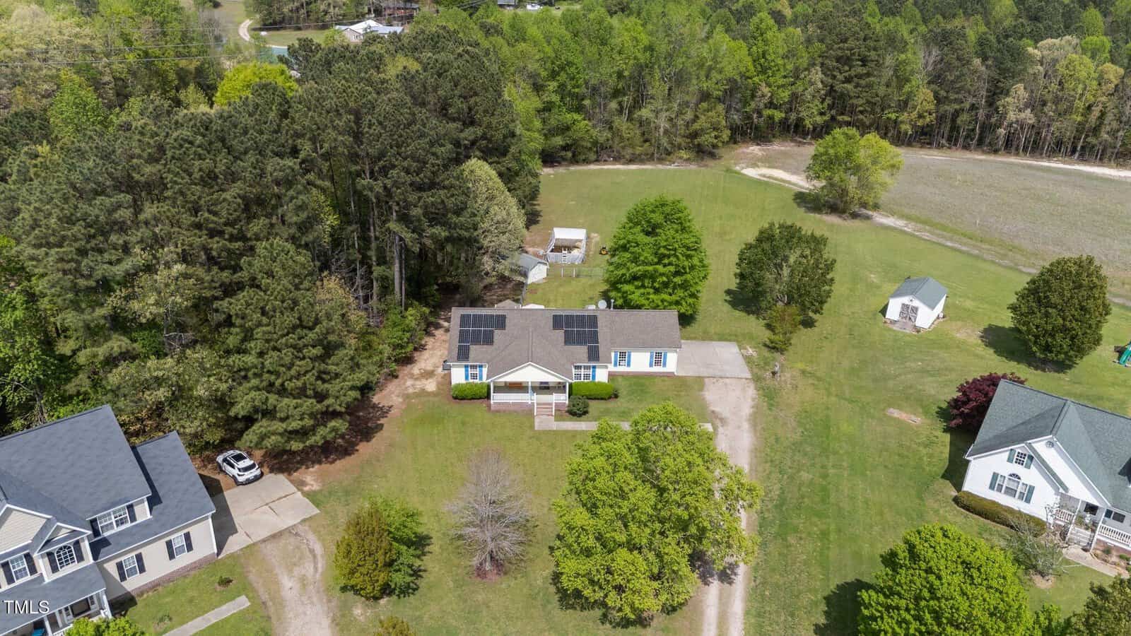 Aerial view of a single-story house with solar panels on the roof, surrounded by trees and grass, with a driveway leading to the house and neighboring properties visible nearby.