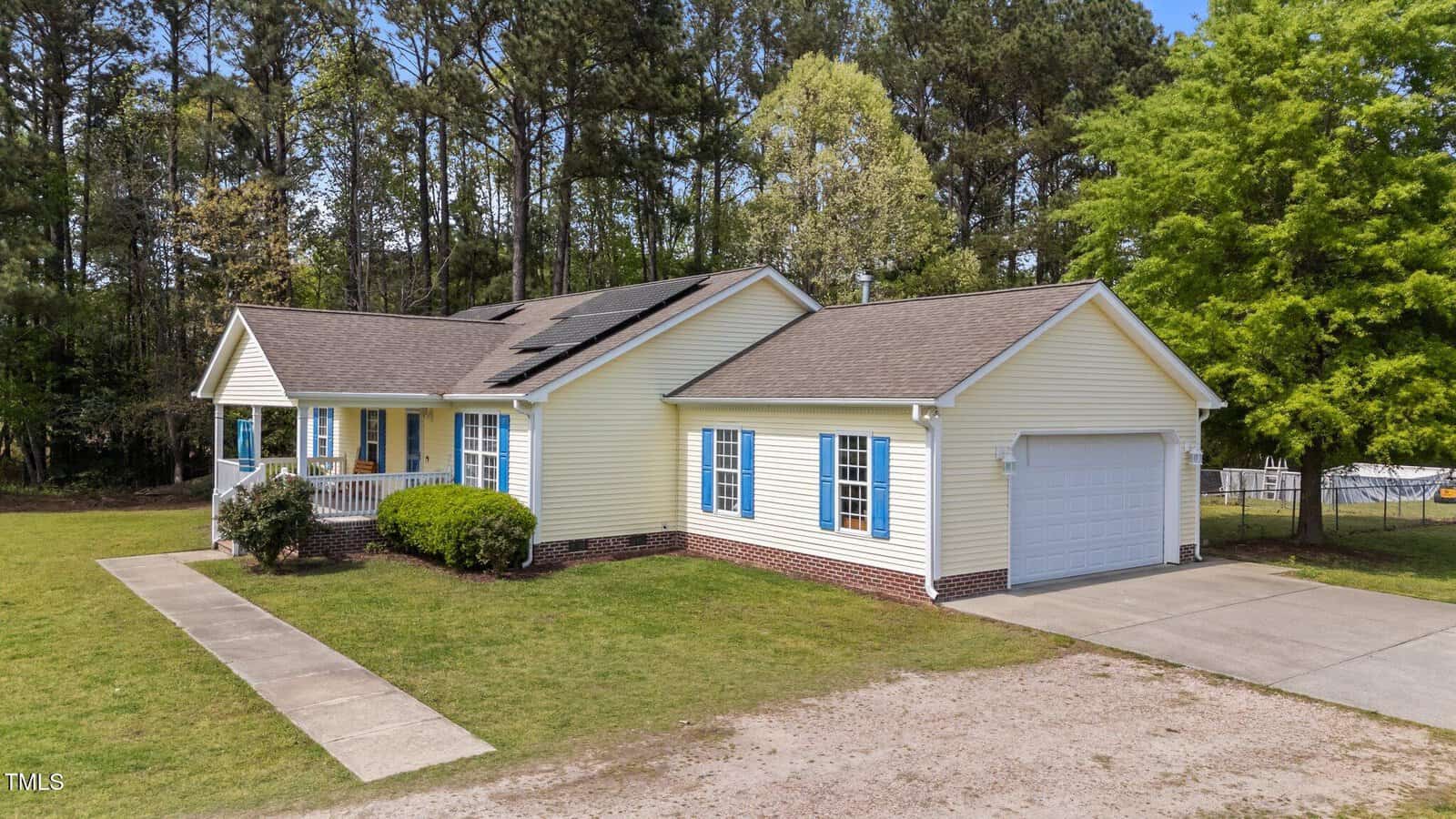 Single-story yellow house with blue shutters, white trim, and a covered front porch. Attached two-car garage, concrete driveway, green lawn, and trees in the background under a clear sky.