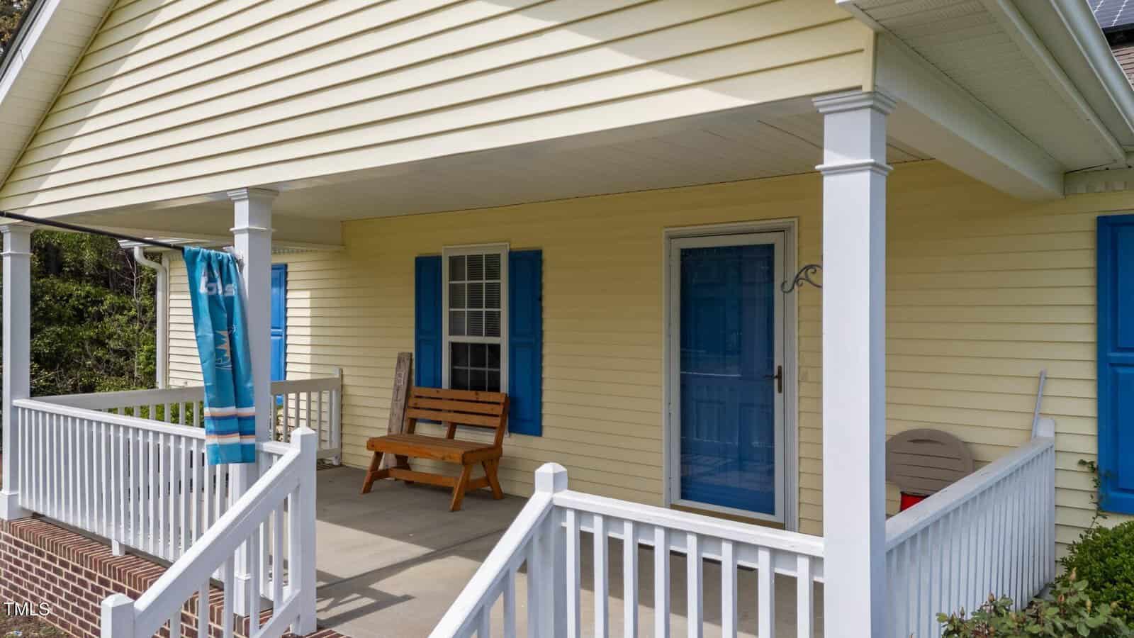 A yellow house with blue shutters and a blue storm door. The front porch has white railings, a wooden bench, a rolled-up outdoor curtain, and two stacked chairs in the corner near the window.