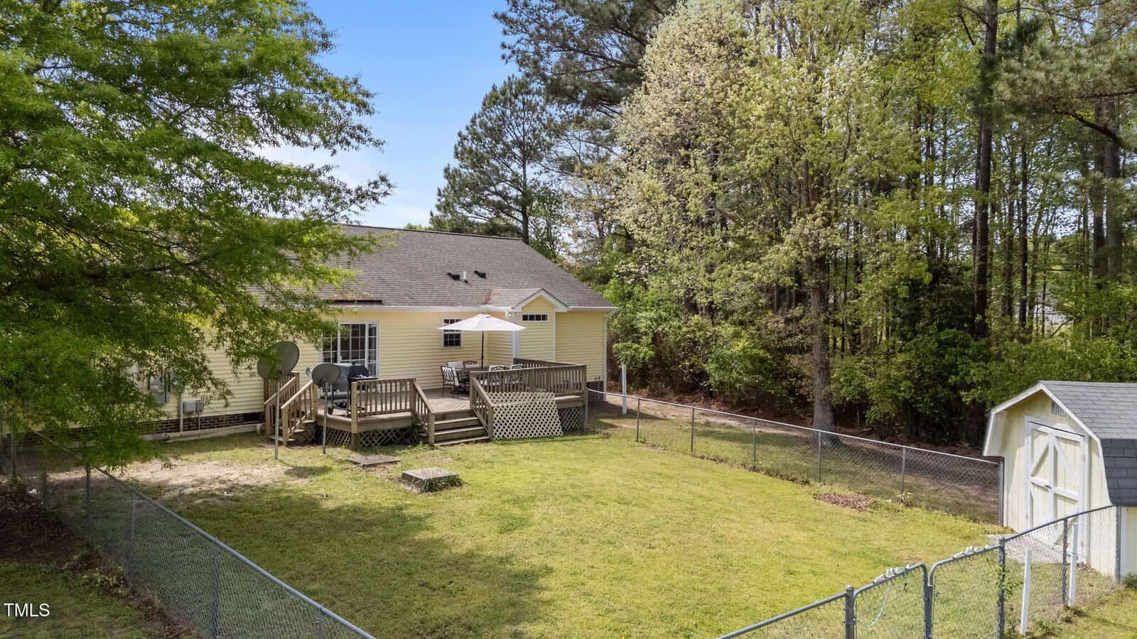 A fenced backyard features a lawn, a wooden deck with patio furniture and a satellite dish, a small shed, and trees surrounding the area. The house exterior is yellow with a sloped roof.