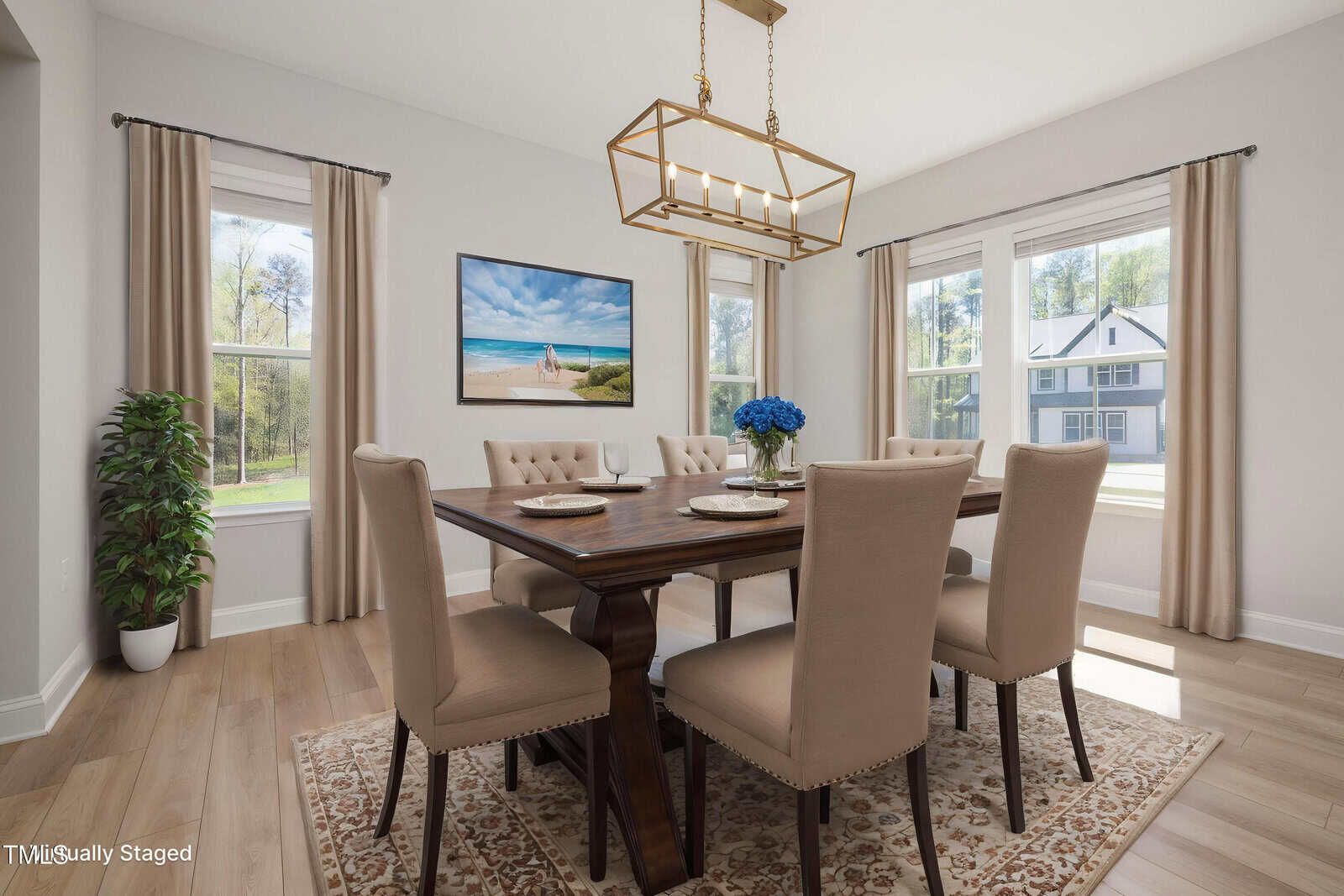 A dining room with a wooden table set for four, beige upholstered chairs, a patterned rug, a gold pendant light, large windows with beige curtains, a potted plant, and a wall picture of a beach scene.