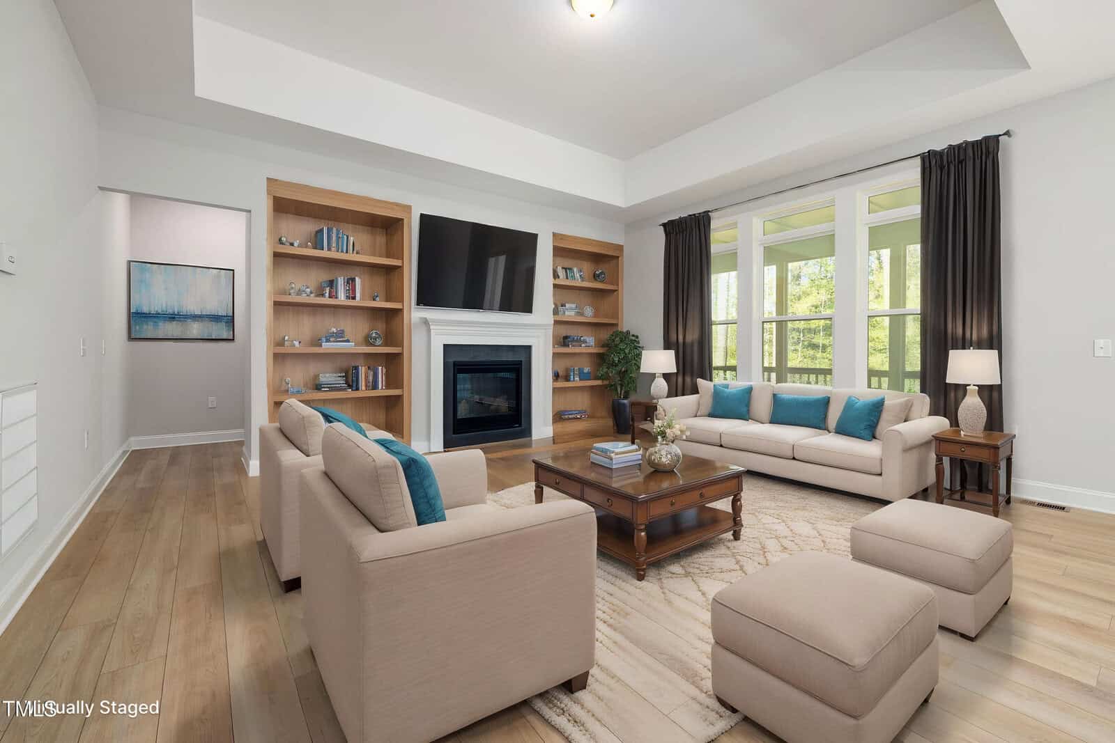 A living room with beige sofas, aqua blue pillows, a wooden coffee table, built-in bookshelves, a fireplace with a mounted TV, large windows with dark curtains, and light wood flooring.