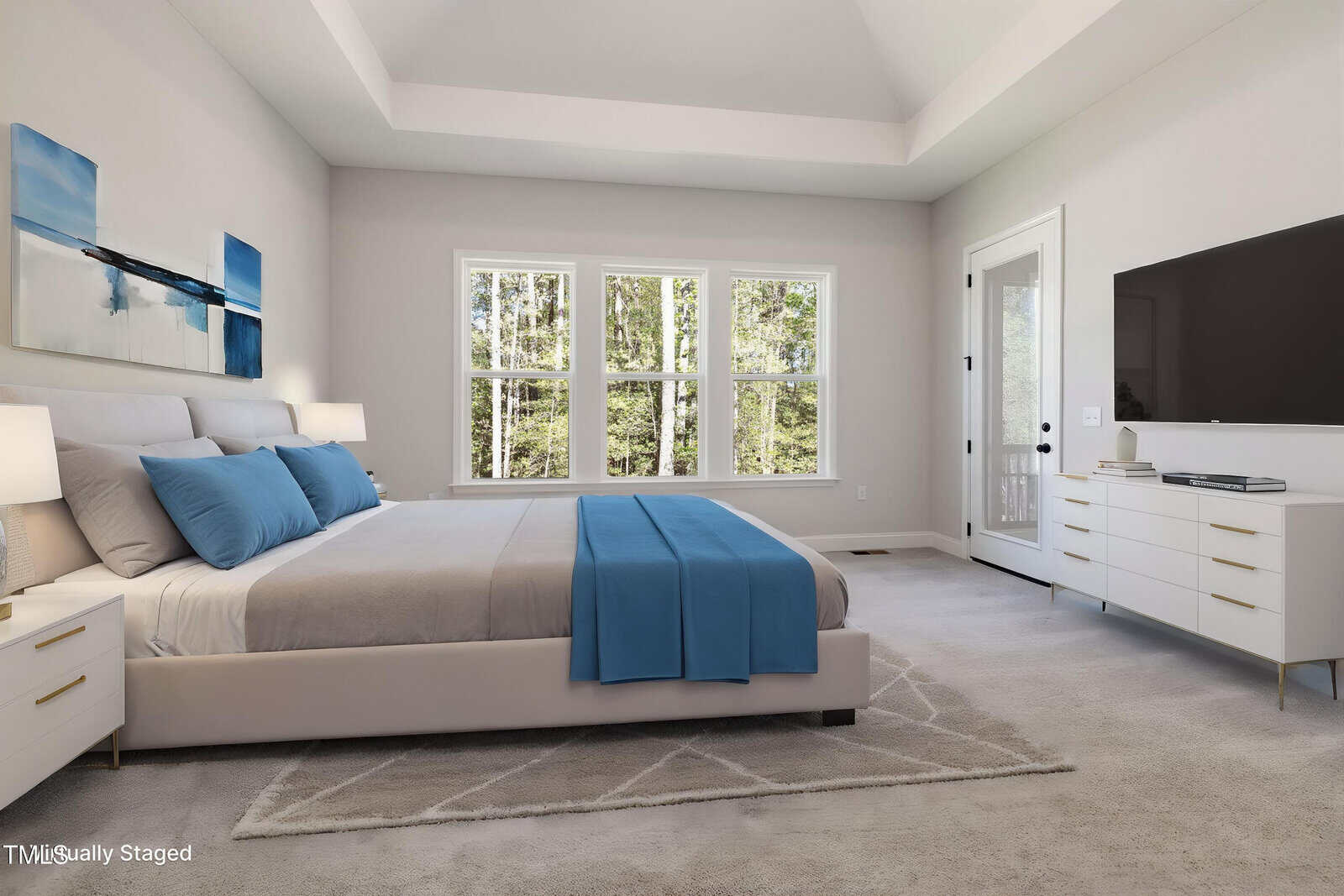 A modern bedroom with a large bed featuring blue and gray bedding, two nightstands with lamps, a white dresser with a flat-screen TV, and three large windows letting in natural light. A glass door is on the right.