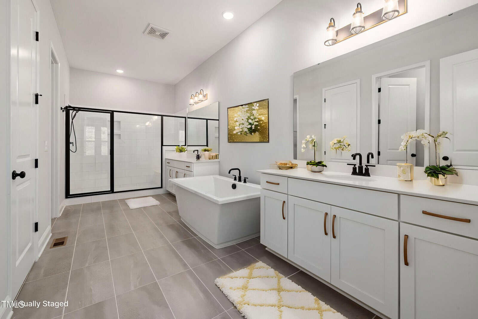 Modern bathroom with a freestanding tub, double sink vanity, large glass-enclosed shower, white cabinets, black fixtures, gray tile floor, and minimal decor including orchids and a wall painting.