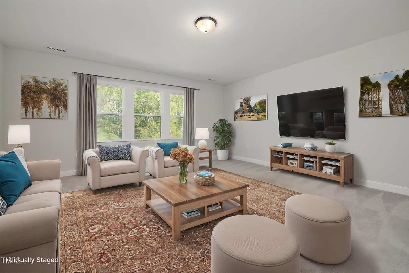 A living room with beige sofas and chairs, a wooden coffee table, a large TV on a wooden console, two round ottomans, lamps, wall art, a patterned rug, and a window with gray curtains letting in natural light.