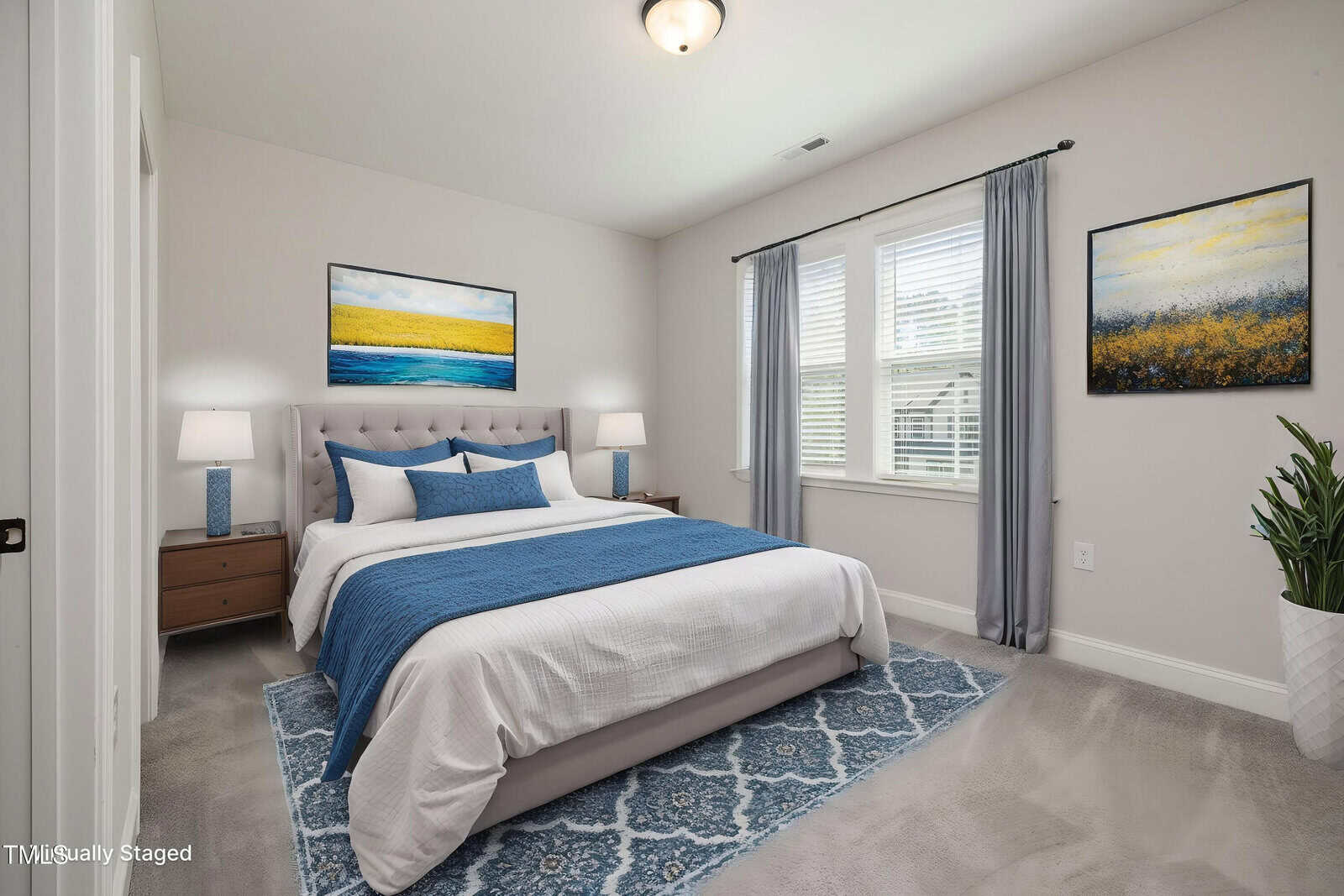A bedroom with a bed featuring white and blue bedding, matching blue lamps on wooden nightstands, two abstract paintings, a blue rug, gray carpet, gray curtains, and a potted plant by a window with blinds.