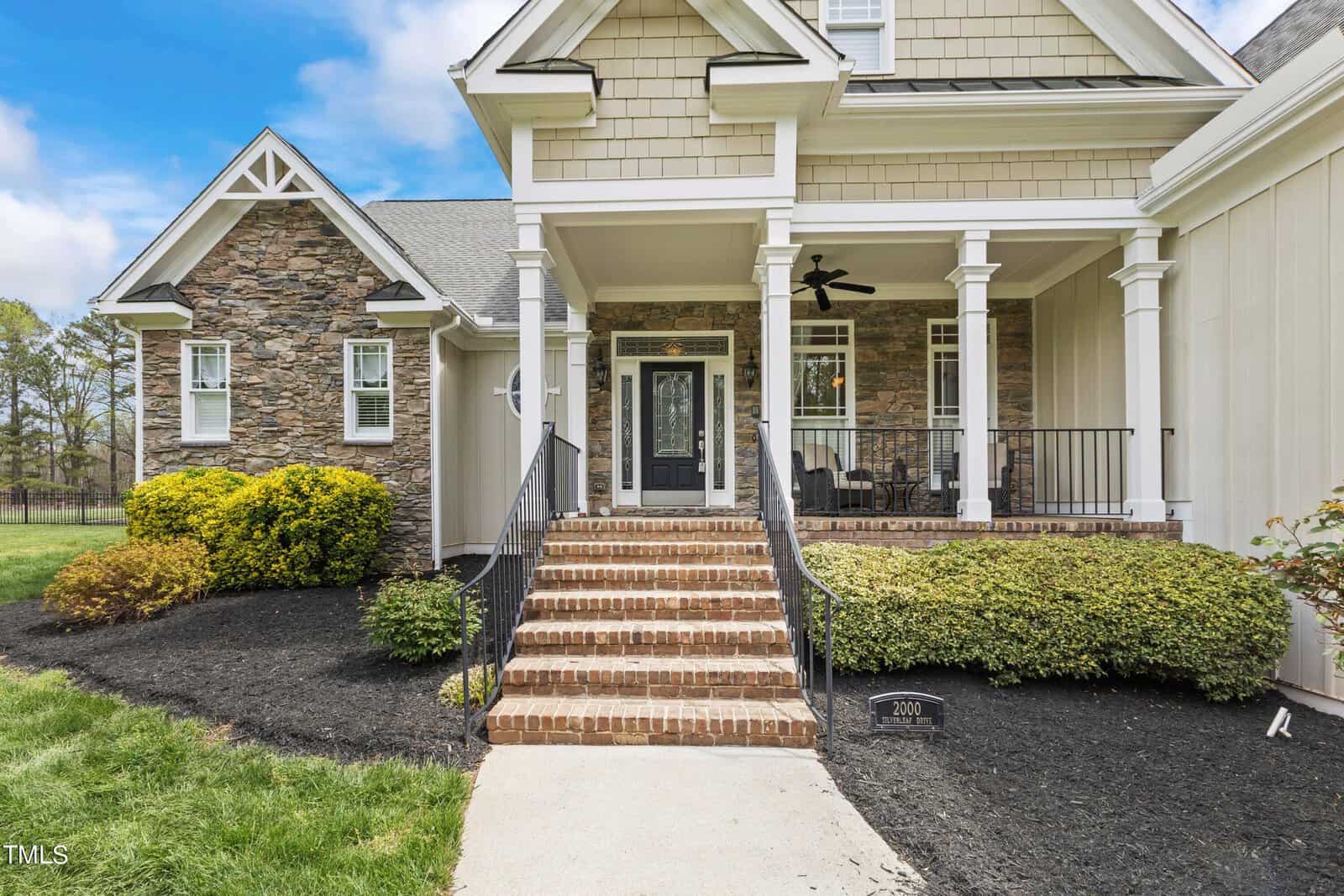 A house with a stone and siding exterior features a front porch with columns and a ceiling fan. Brick steps lead up to a black front door with decorative glass. Bushes are planted around the walkway and a lawn is visible in the foreground.