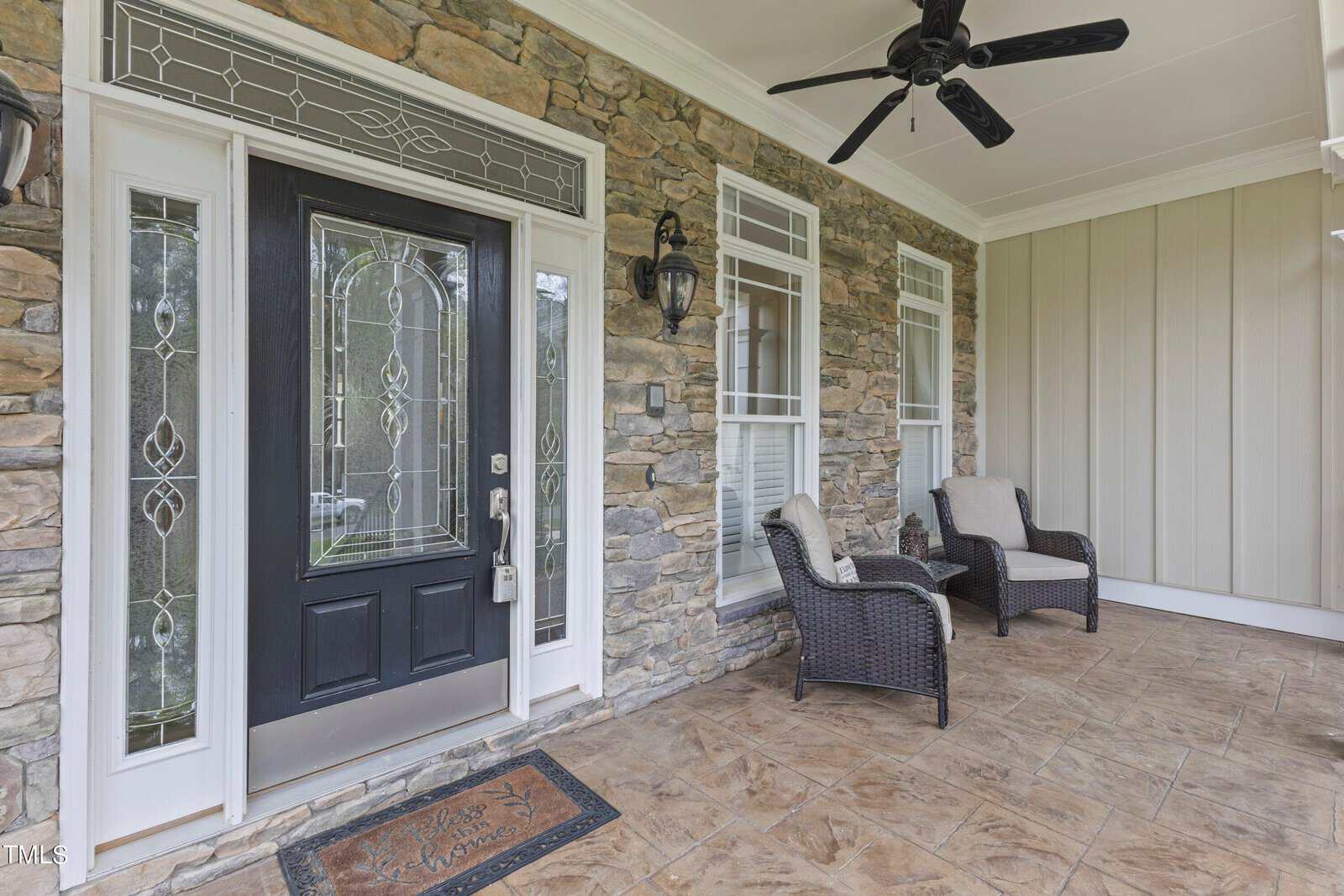 A porch with a stone wall features a black front door with decorative glass panels and two small windows. Two wicker chairs with cushions are placed on the tile floor. A ceiling fan hangs above, and wall-mounted lanterns flank the windows.