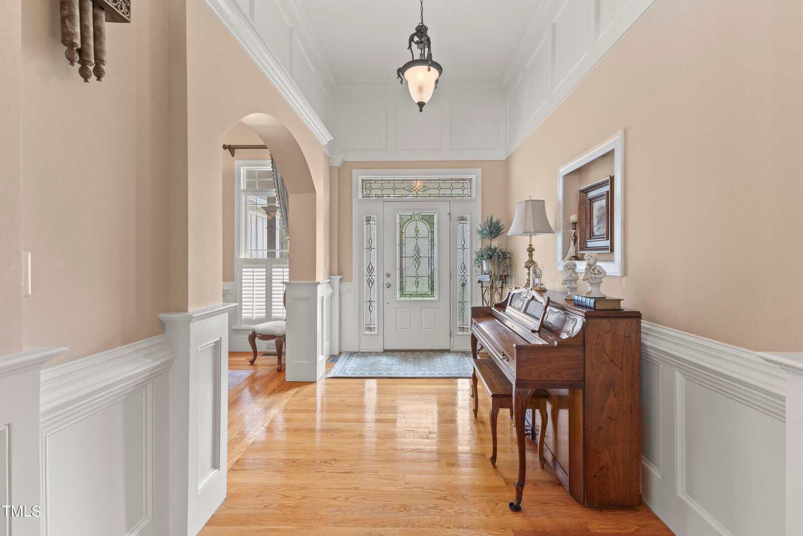 A hallway with wooden floors, white wainscoting, and light-colored walls. A piano with a lampshade and framed artwork sits on the right. An arched opening leads to another room. A front door with decorative glass panels is visible at the end.