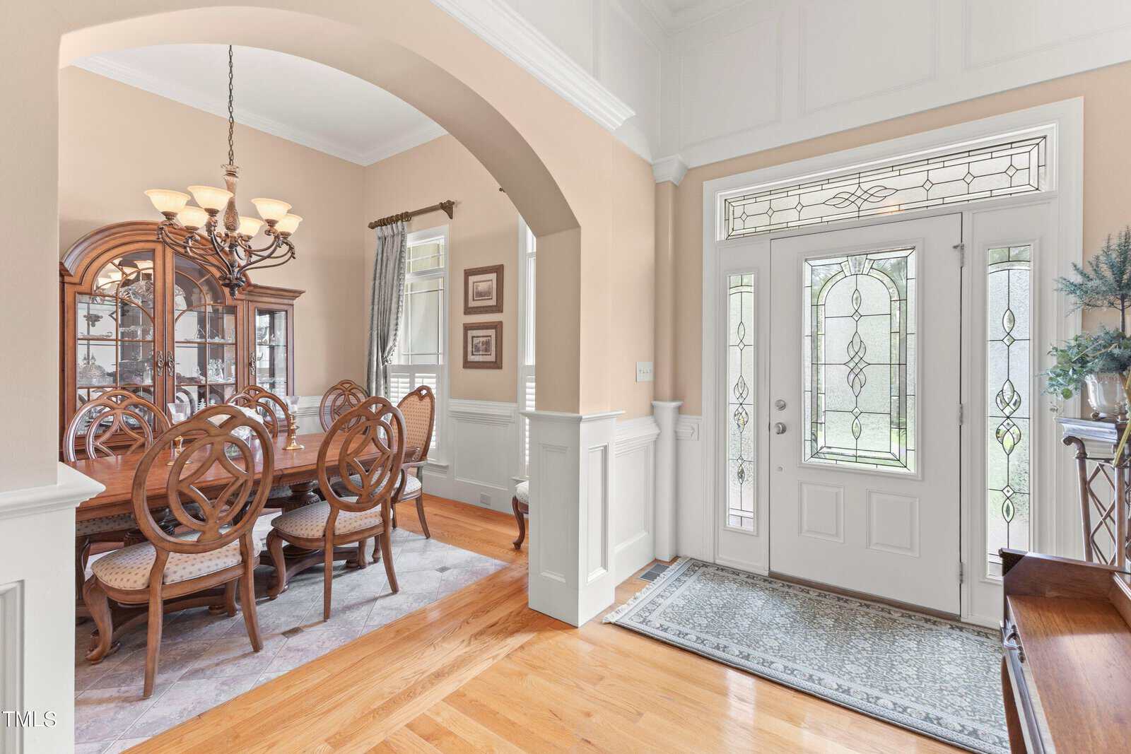 Bright dining room with a wooden table and eight chairs, lit by a chandelier. The room features arched entryways and beige walls. A large windowed front door is visible, alongside a small table with a plant.
