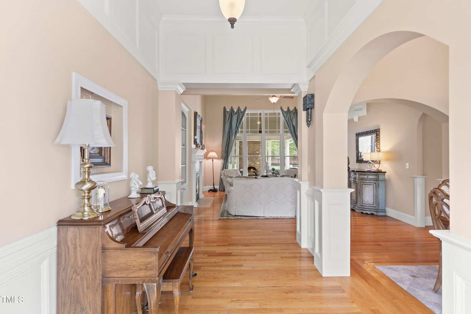 A living room with light-colored walls, hardwood floors, and a wooden piano on the left. It features a sofa, a lamp, and curtains in the background. Arched doorways and framed artwork add to the decor.