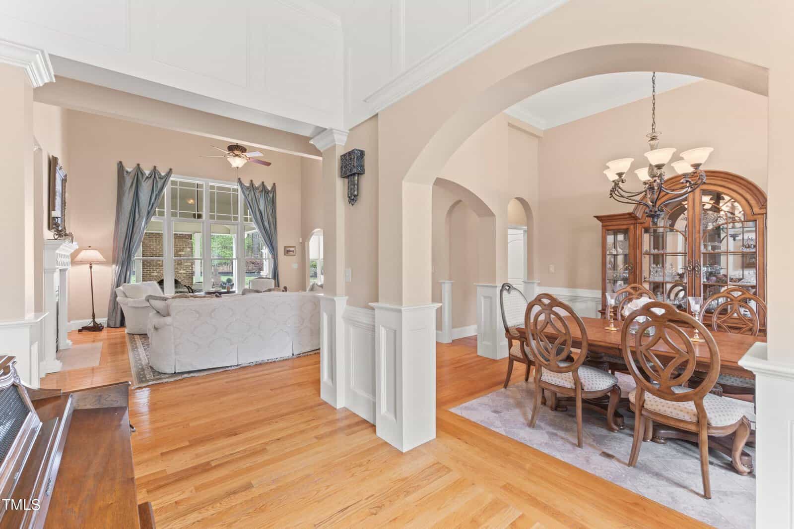 A spacious open-concept living and dining area with hardwood floors. The dining room features a wooden table and chairs under a chandelier. The living room has a couch and a large window with curtains. Both rooms are connected by arches and columns.