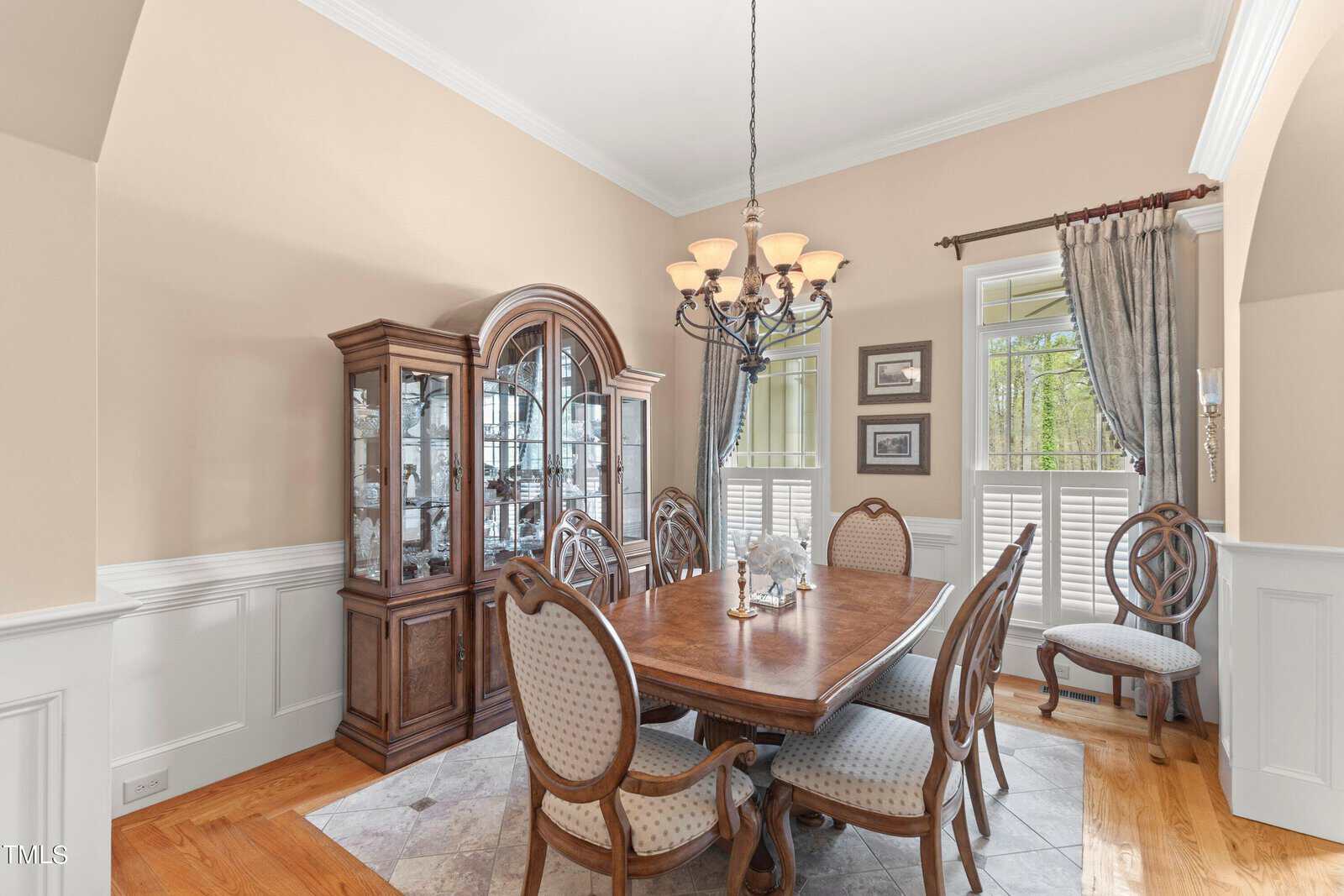 A dining room with a wooden table and six upholstered chairs. A wooden china cabinet is against the wall. A chandelier hangs above the table. Windows on the right have curtains and shutters. The room has light-colored walls and a wooden floor.