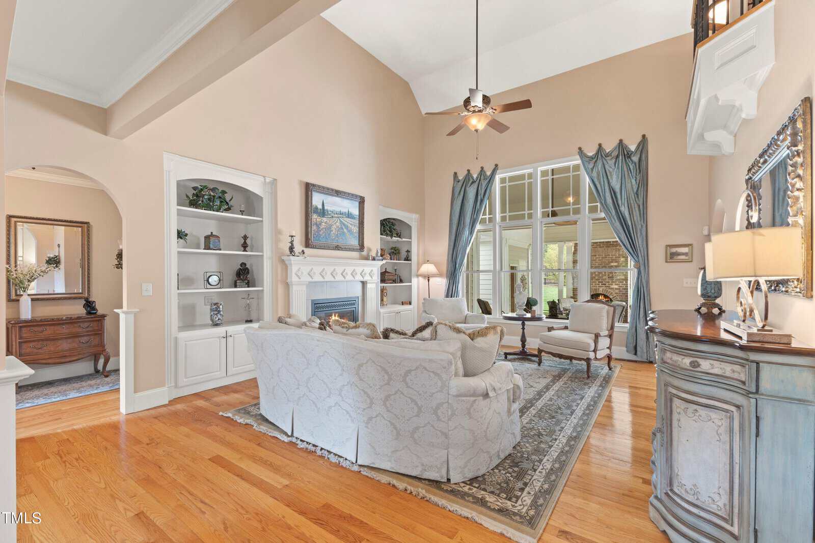 A spacious living room with hardwood floors features a sitting area with sofas and armchairs around a coffee table. Large windows with blue curtains allow natural light. A ceiling fan hangs from the high ceiling, and a fireplace is on the side wall.