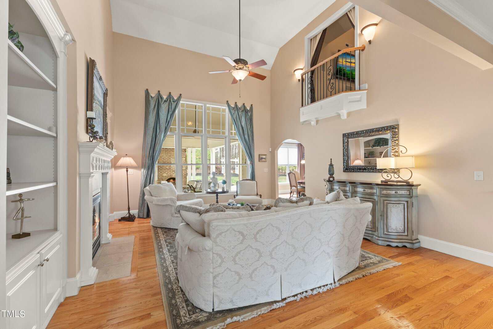 A spacious living room with high ceilings, featuring a matching set of patterned sofas and armchairs around a rug. There's a fireplace, built-in shelving, and large windows. A ceiling fan is overhead, and stairs lead to a small upstairs balcony.