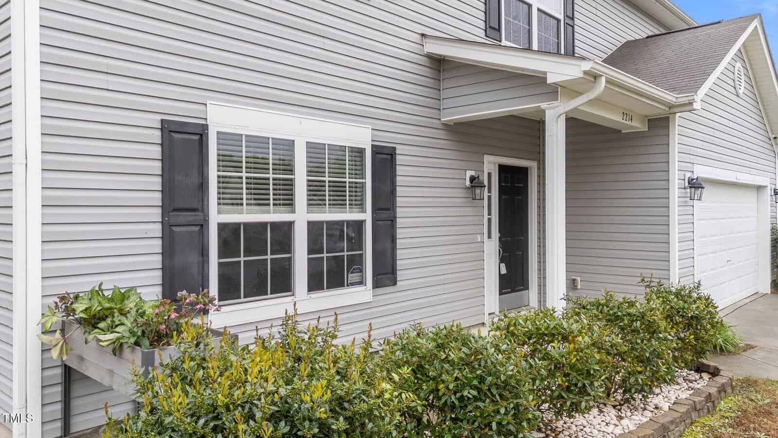 Exterior of a modern house with light gray siding and white trim. There is a black door with a small overhang, two windows with black shutters, and a garage on the right. Shrubs and potted plants line the front of the house.
