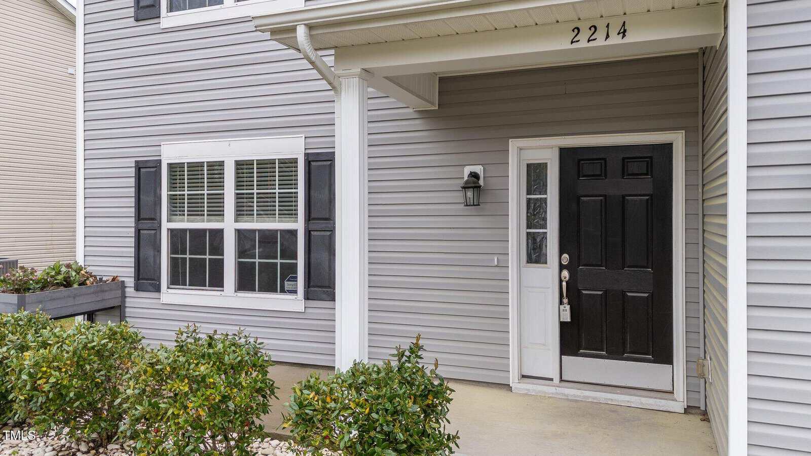 A gray house exterior with a white-trimmed window and a black front door. The address "2214" is displayed above the door. A small porch light is mounted next to the door. Bushes line the front of the house.