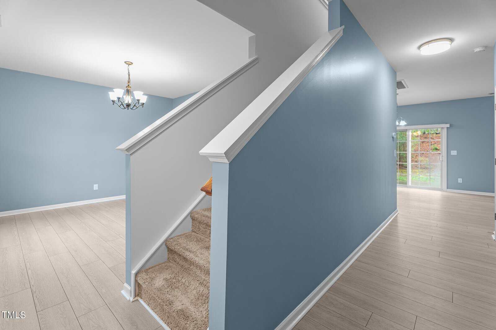 Interior of a house featuring a staircase with beige carpet and blue walls. The area has light wood flooring, a ceiling light fixture, and a sliding glass door leading to a garden outside.