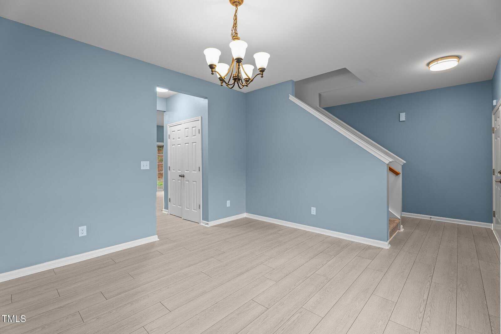 A room with light blue walls and light wood flooring features a white staircase to the right. A white door opens to another space. A gold and white chandelier hangs from the ceiling, and a circular ceiling light is near the stairs.